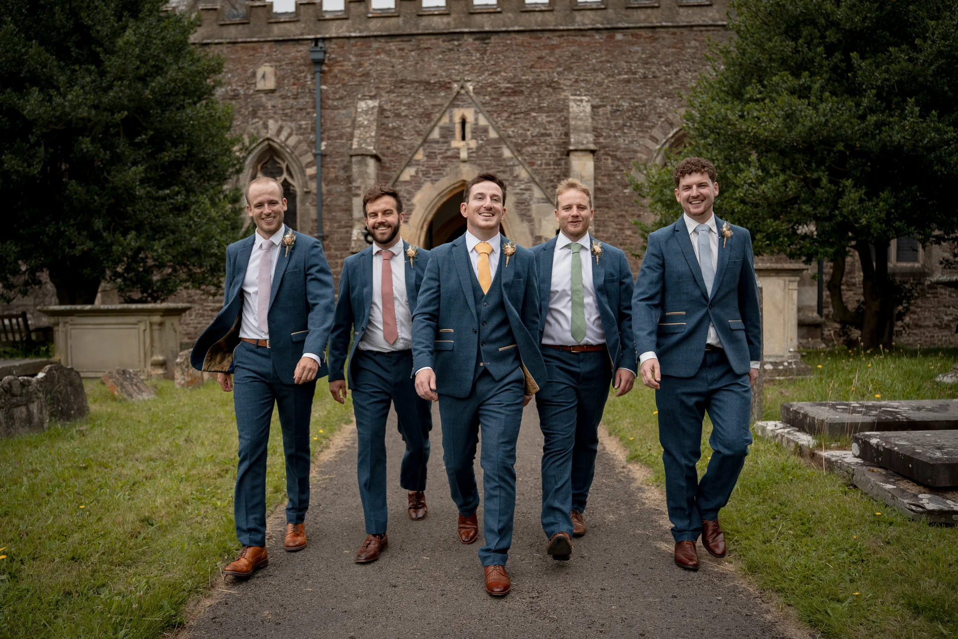 Five smiling groomsmen in blue suits walk together along a stone path outside an ancient stone church.