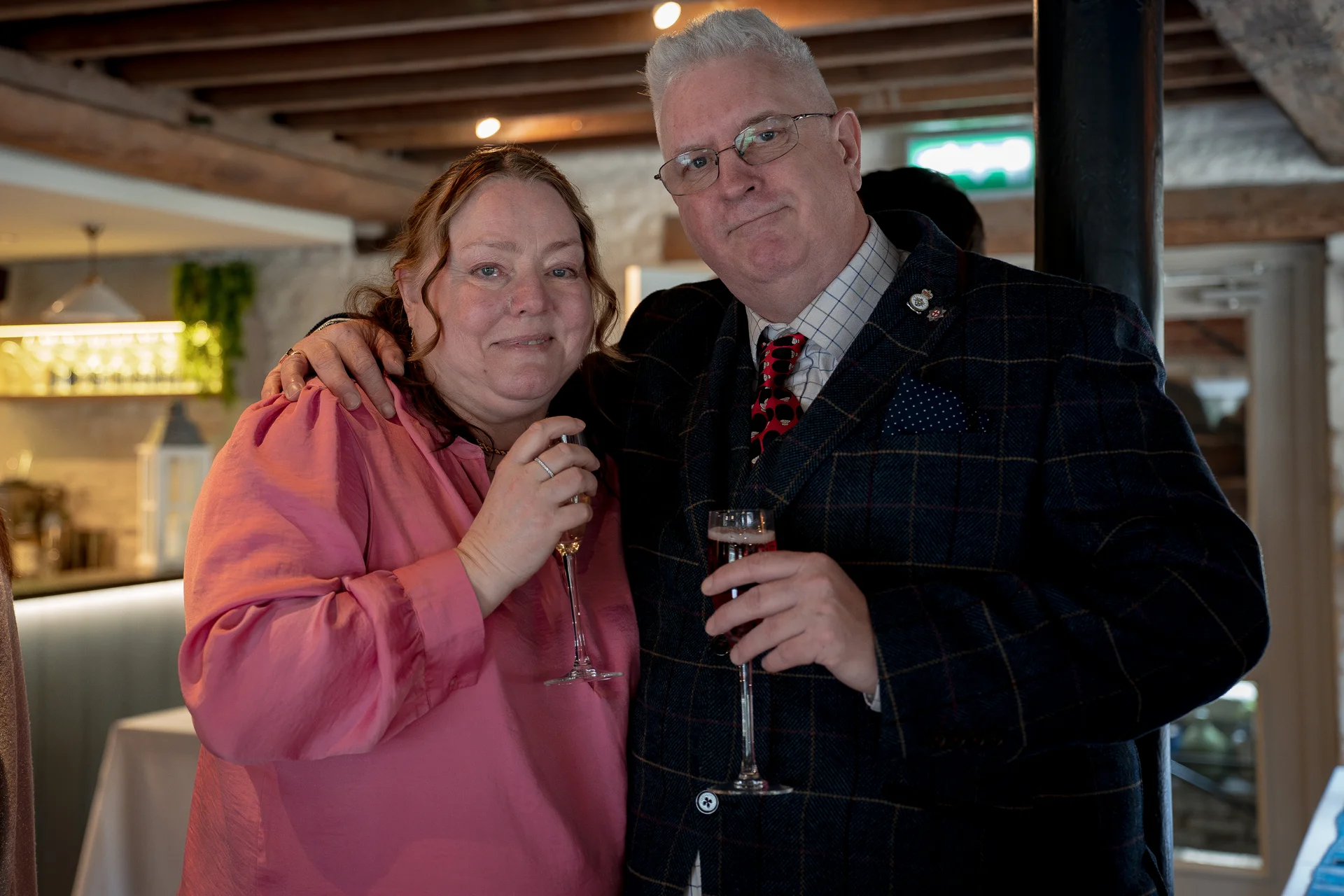An older couple stand together smiling at a wedding reception inside a rustic venue with exposed wooden rafters, each holding a glass of champagne.
