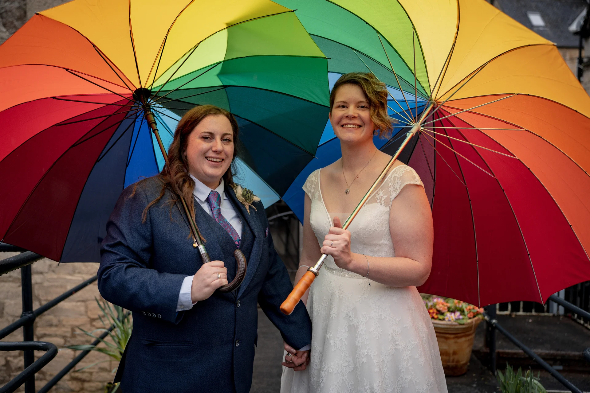 A newlywed same-sex couple proudly hold large rainbow umbrellas together outdoors while smiling and holding hands.