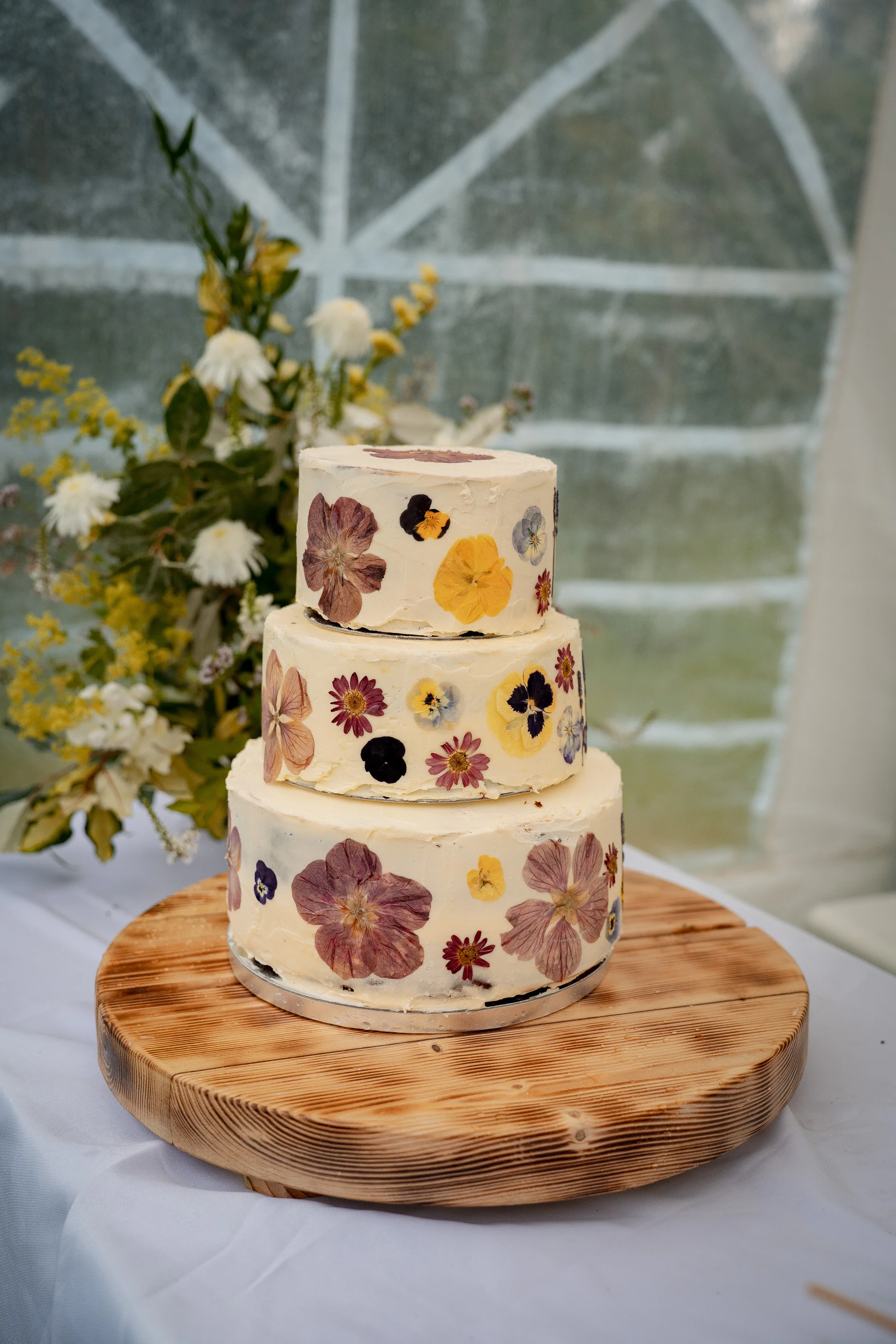 A beautiful three-tiered wedding cake decorated with pressed edible flowers sits proudly on a rustic wooden stand against a backdrop of soft white florals and natural light.