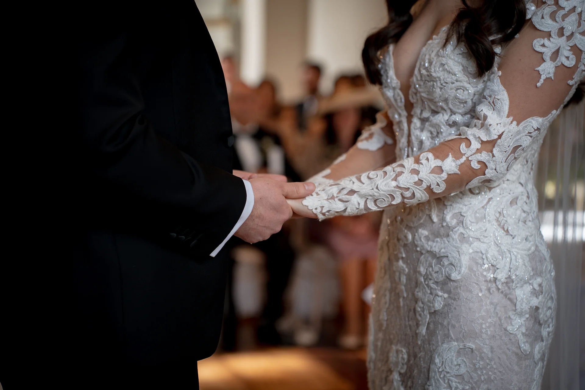 A bride in an intricate lace long-sleeve gown and a groom in a black tuxedo hold hands during their wedding ceremony.