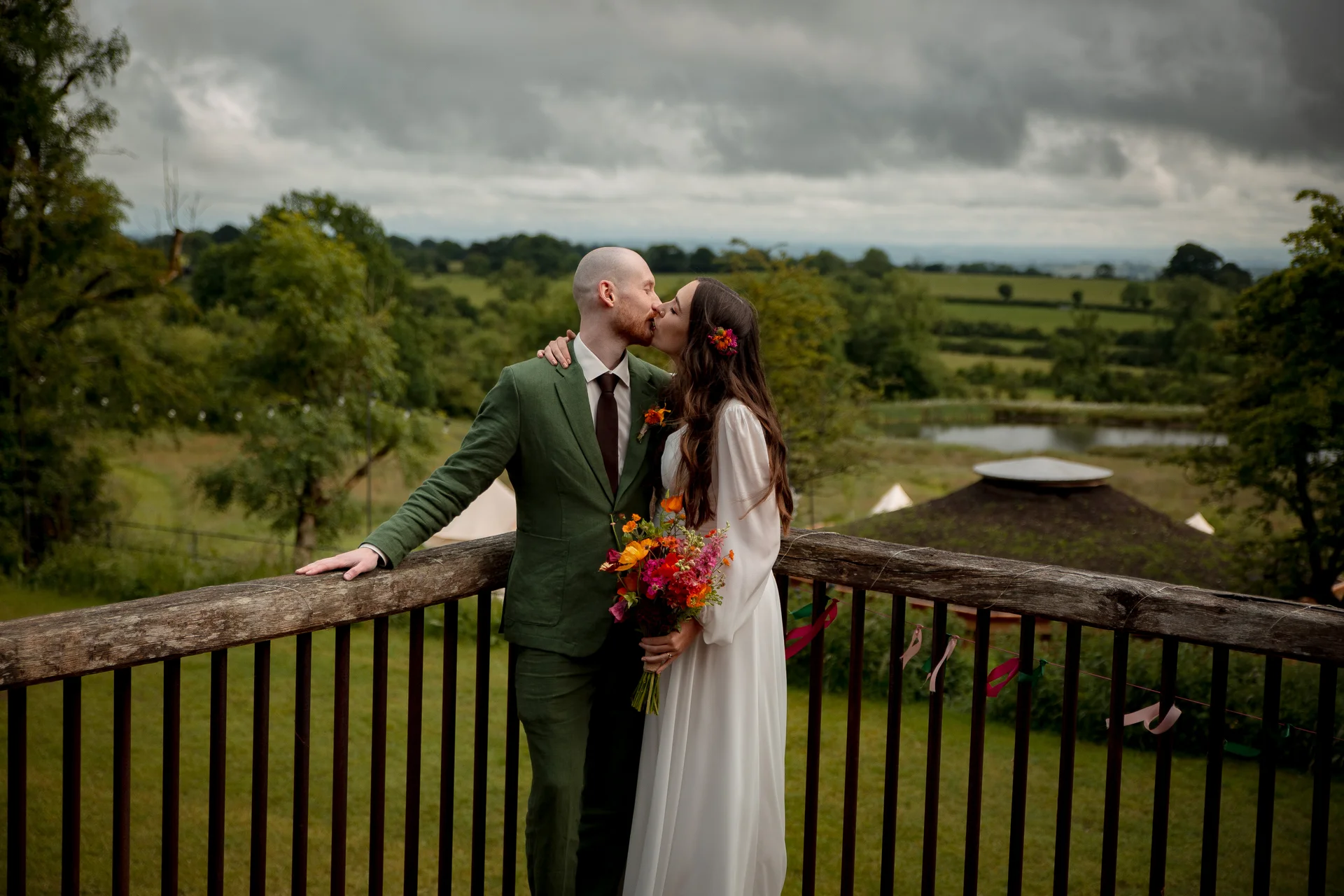 A newlywed couple shares a tender kiss on a wooden balcony overlooking a scenic landscape with rolling hills and a moody sky.