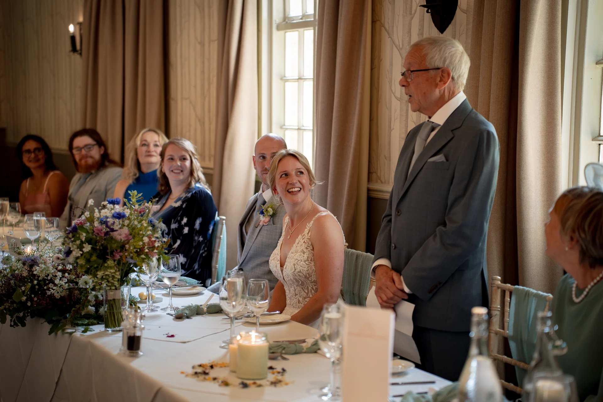 An older man in a grey suit delivers a speech at the top table while the smiling bride and guests look on during the wedding reception.