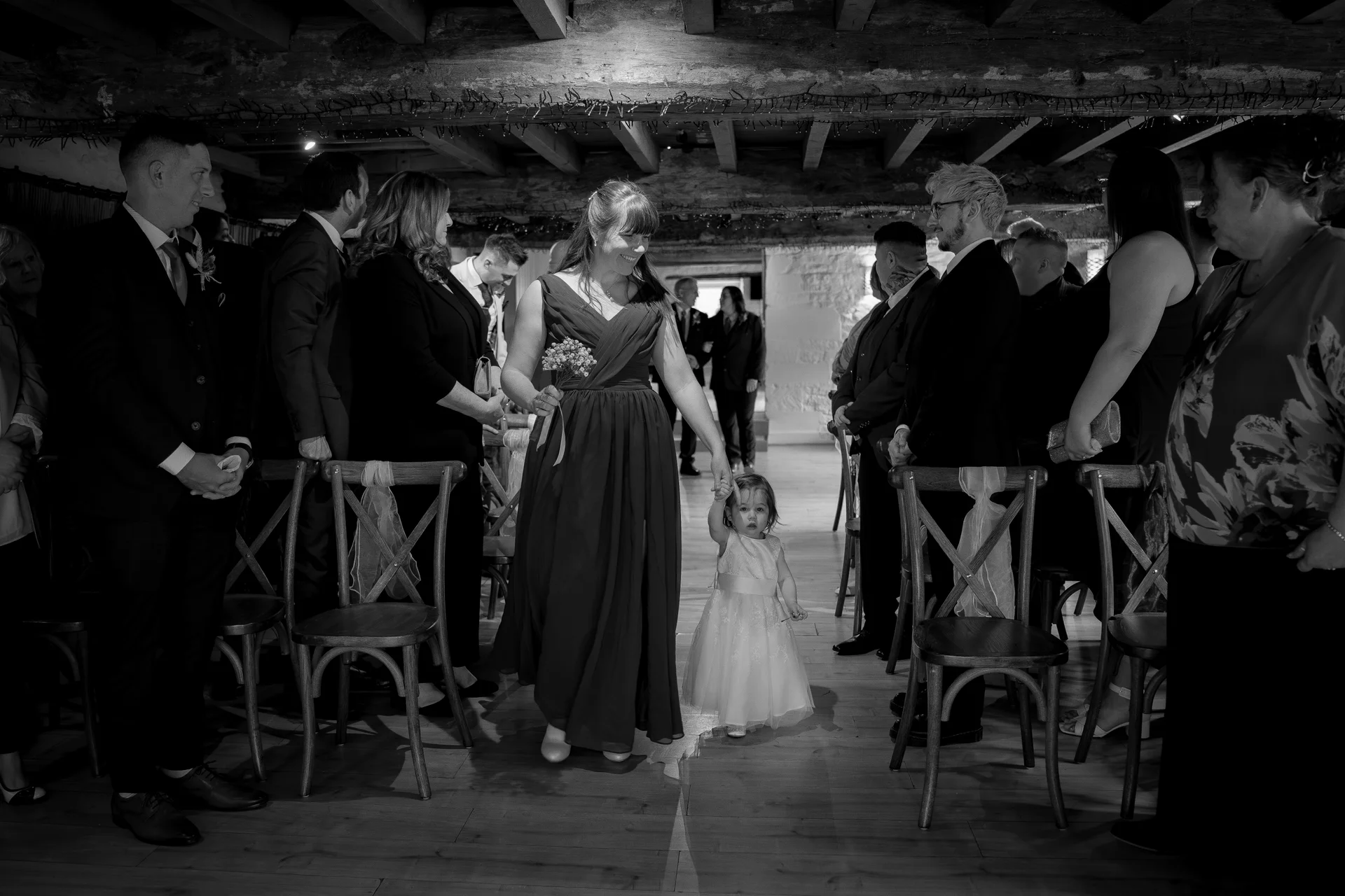 A bridesmaid smiles as she walks down the aisle hand-in-hand with a young flower girl inside a rustic barn wedding venue.