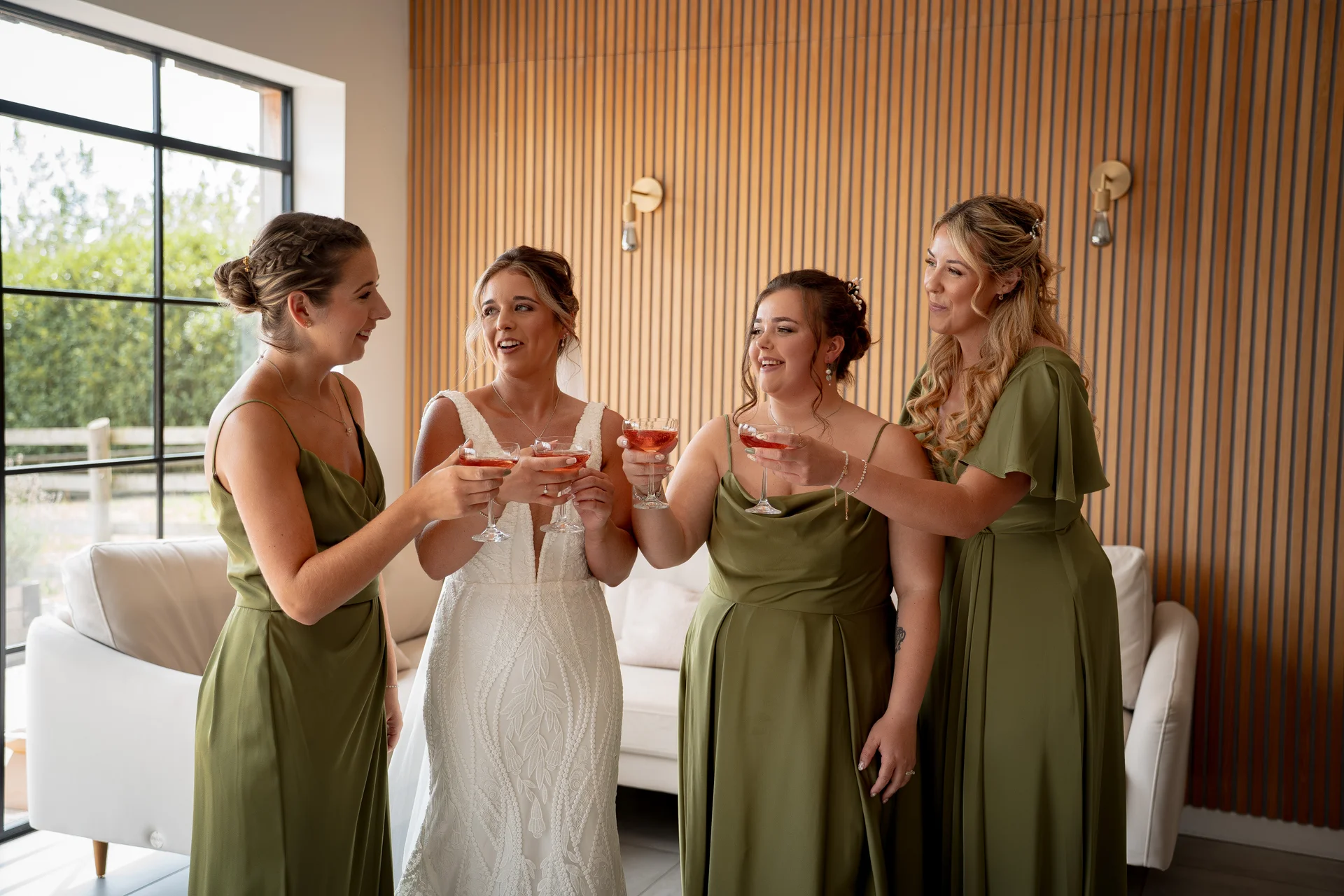 A glowing bride in a lace gown and her three bridesmaids in sage green dresses raise a toast with pink cocktails in a bright, modern room.