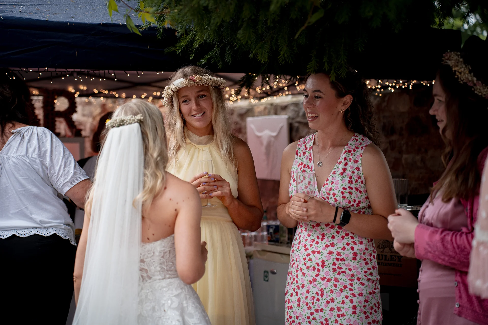 A bride wearing a veil and lace gown chats and shares a joyful moment with her bridesmaids beneath a festoon-lit marquee during a wedding drinks reception.