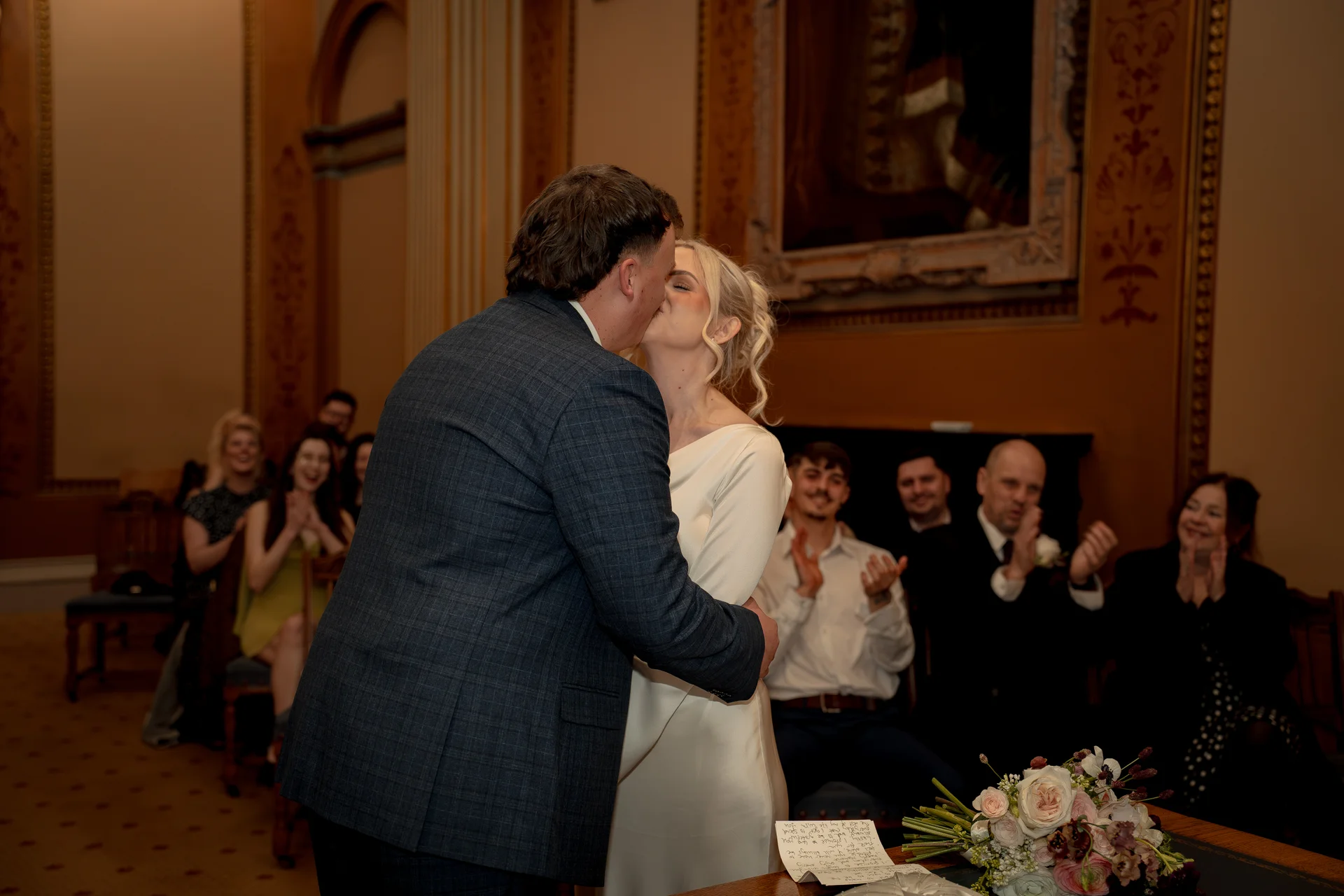 Newlyweds share their first kiss at the altar during a civil ceremony in an elegant stately home, with guests applauding in the background.