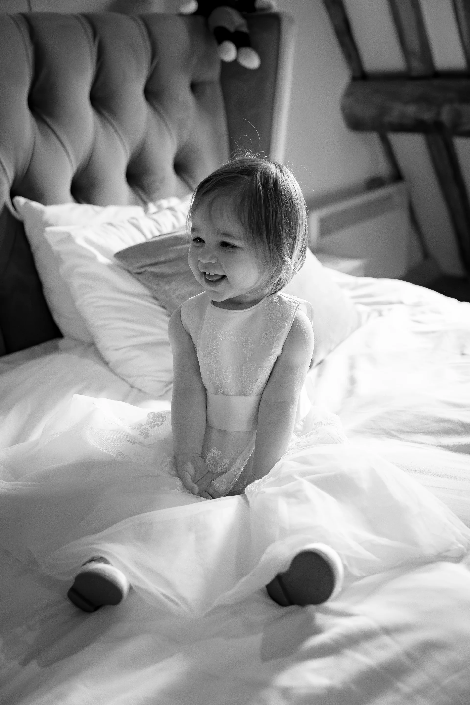 A young flower girl laughs joyfully while sitting on a large white bed during wedding morning preparations.