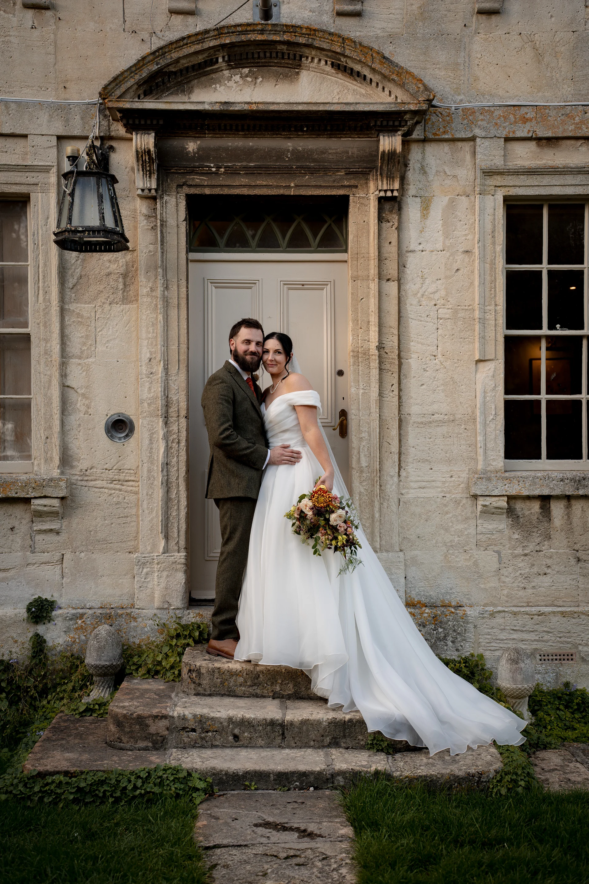 A newlywed couple poses together on stone steps in front of a historic stone doorway of a country house, with the bride holding a colourful autumn bouquet.