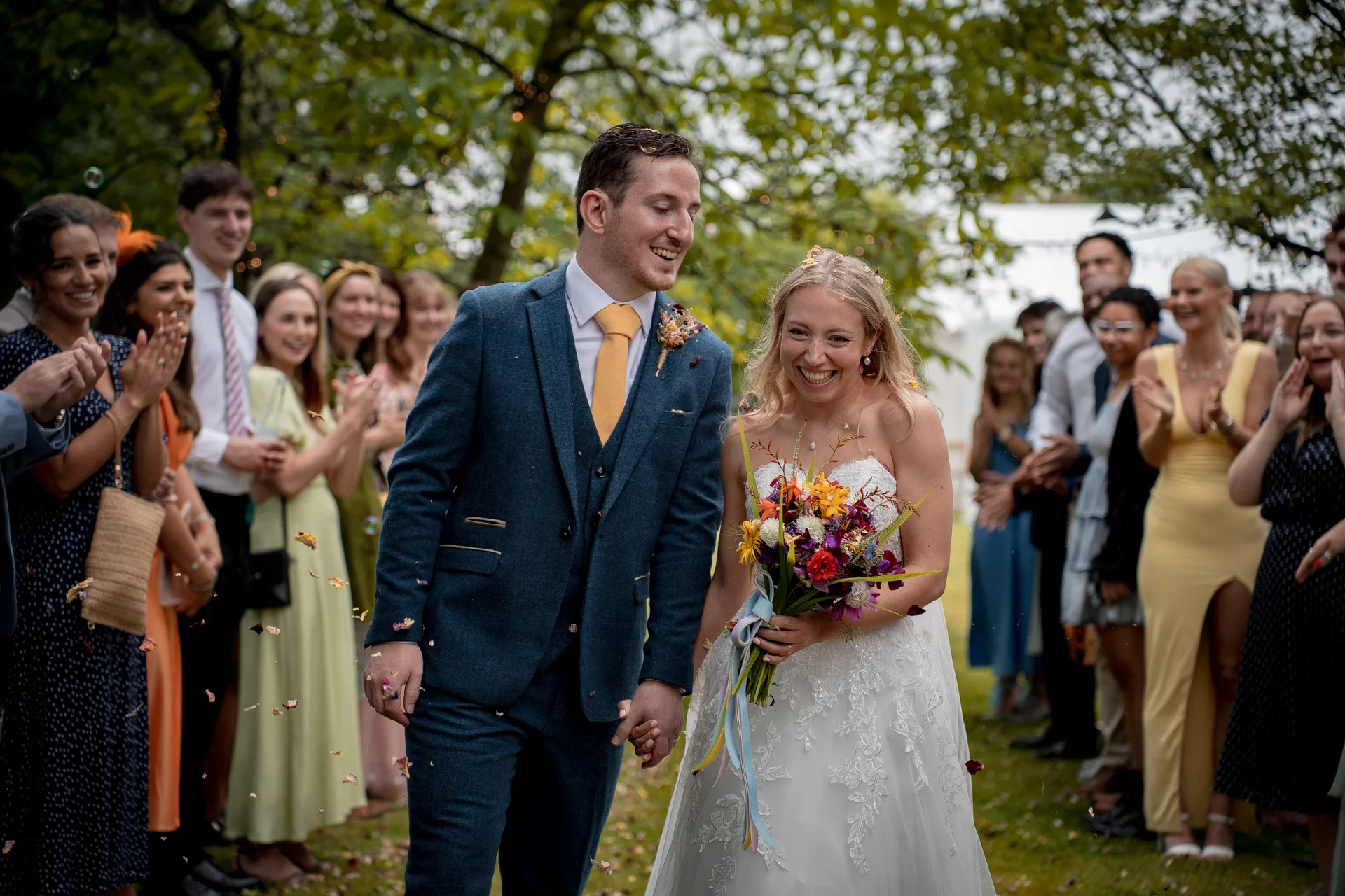 A smiling bride and groom walk hand in hand through a cheering crowd of wedding guests as colorful confetti falls around them in an outdoor garden setting.