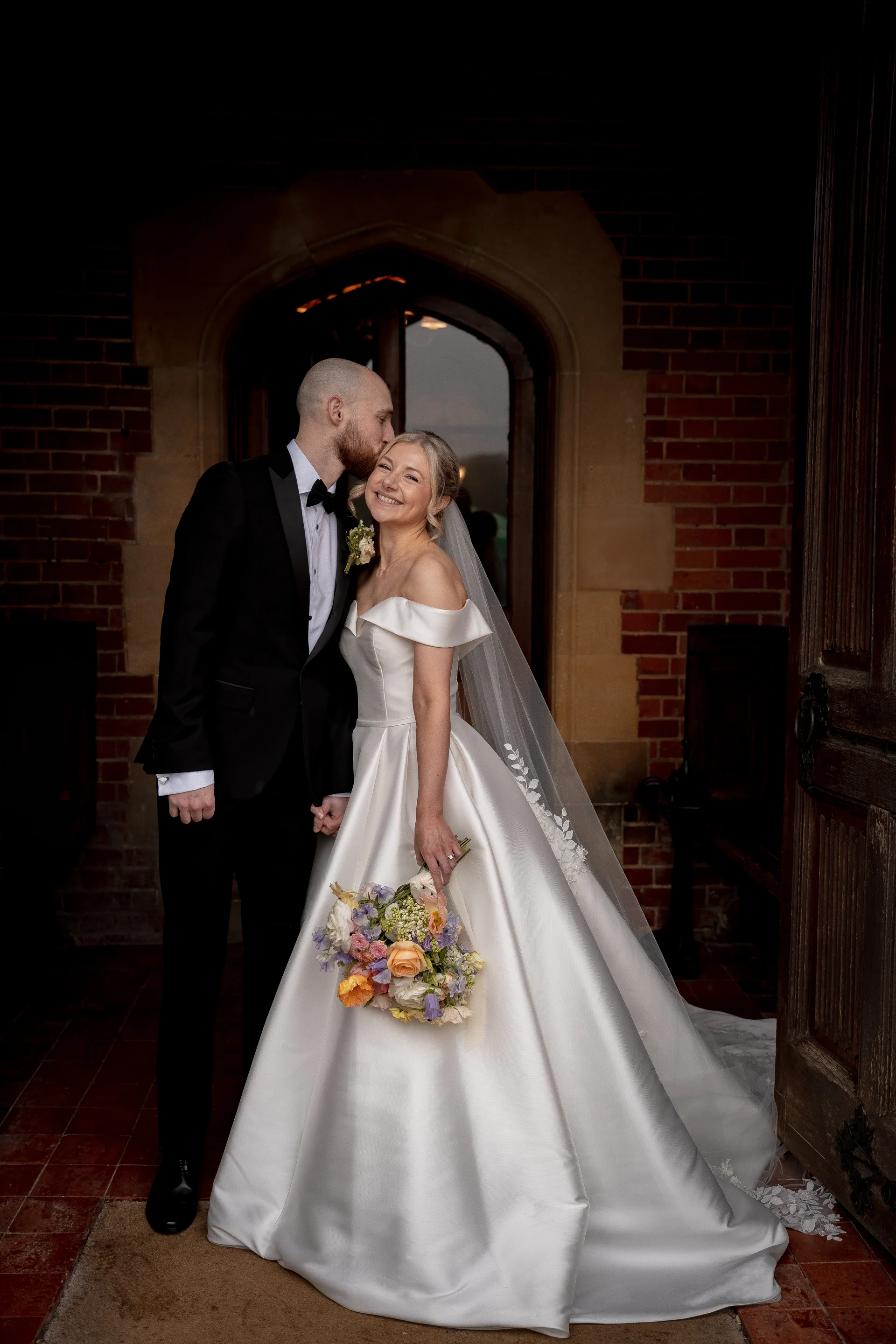 A groom in a classic black tuxedo kisses his smiling bride on the forehead as she holds a colourful bouquet in front of a stone archway and brick doorway.