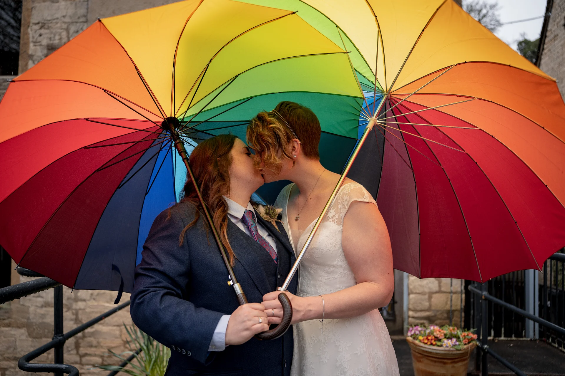 A newlywed same-sex couple shares a tender kiss underneath a vibrant rainbow umbrella on a rainy wedding day.