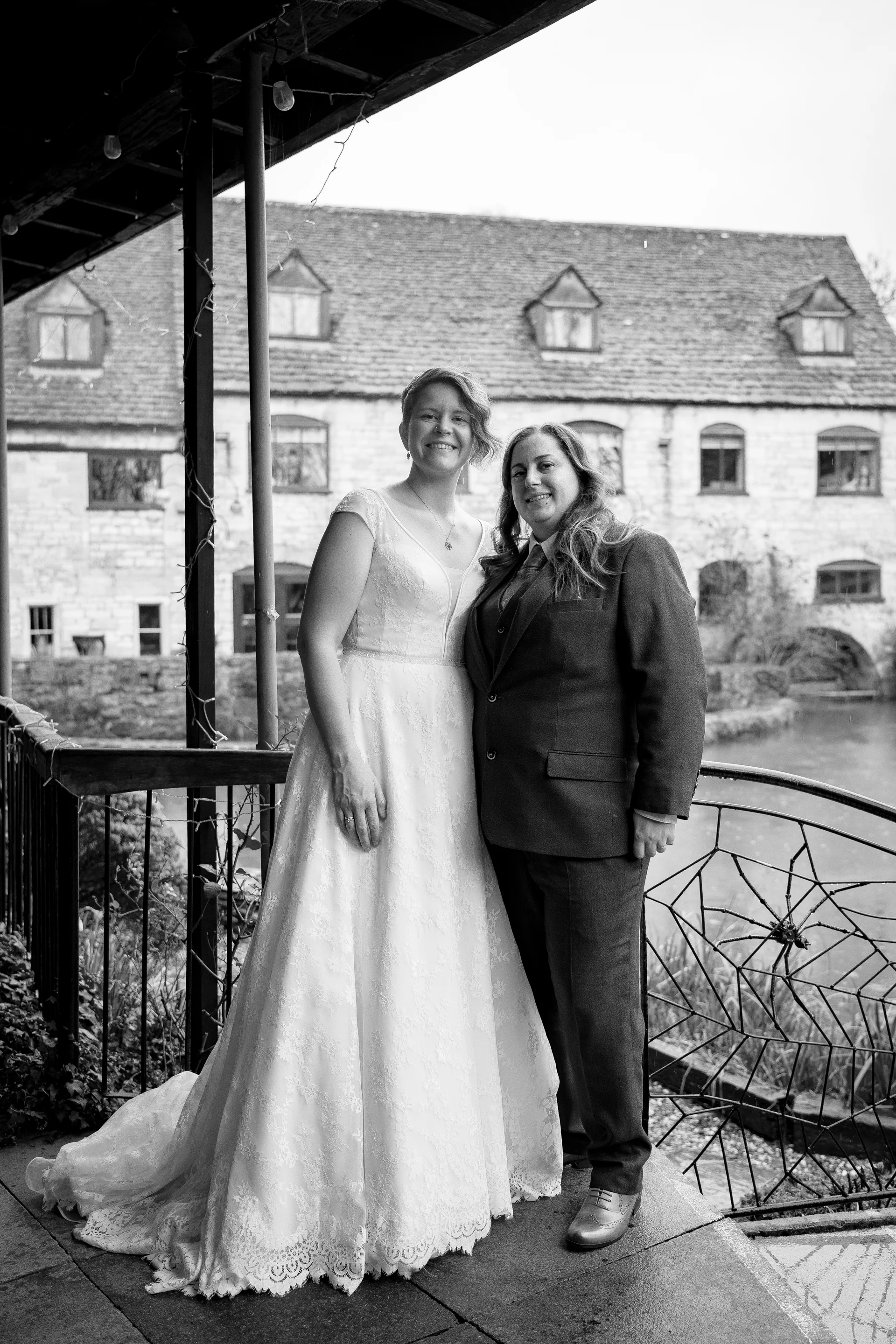 A smiling same-sex couple poses together outdoors on a stone terrace in front of a rustic building, with one bride in a lace A-line gown and the other in a tailored suit.