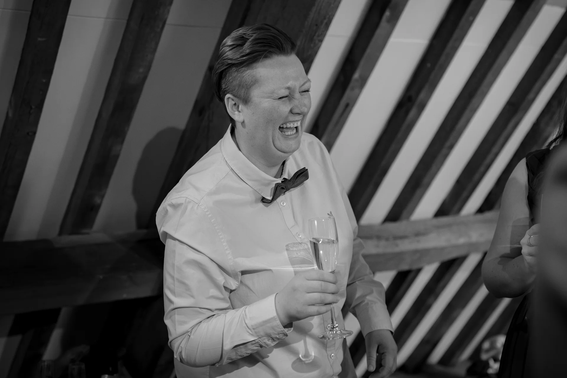 A guest in a collared shirt and bow tie laughs heartily while holding a glass of champagne inside a rustic barn with exposed rafters.