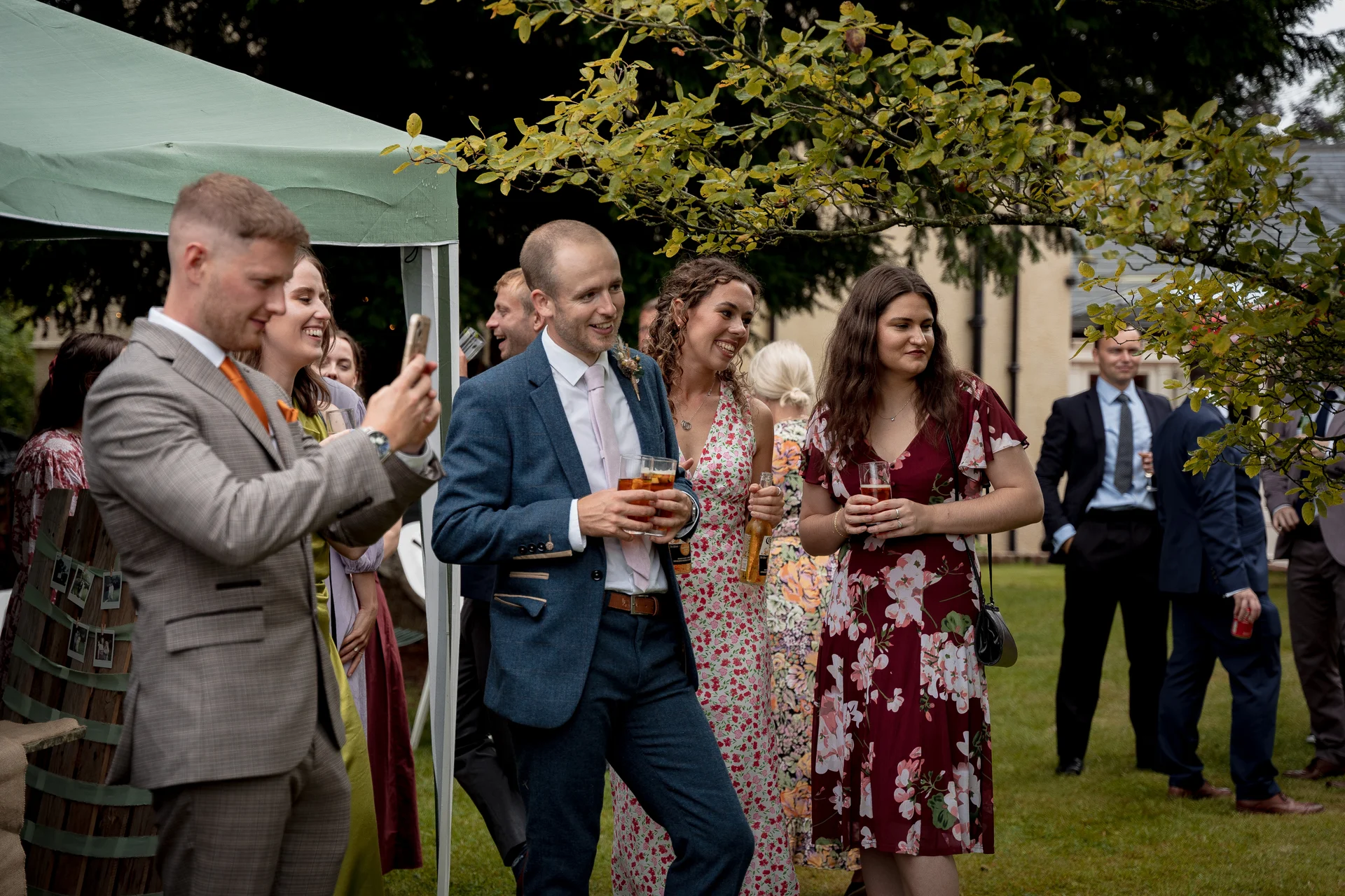 A group of smartly dressed wedding guests mingle and chat happily outdoors during a garden drinks reception near a marquee.