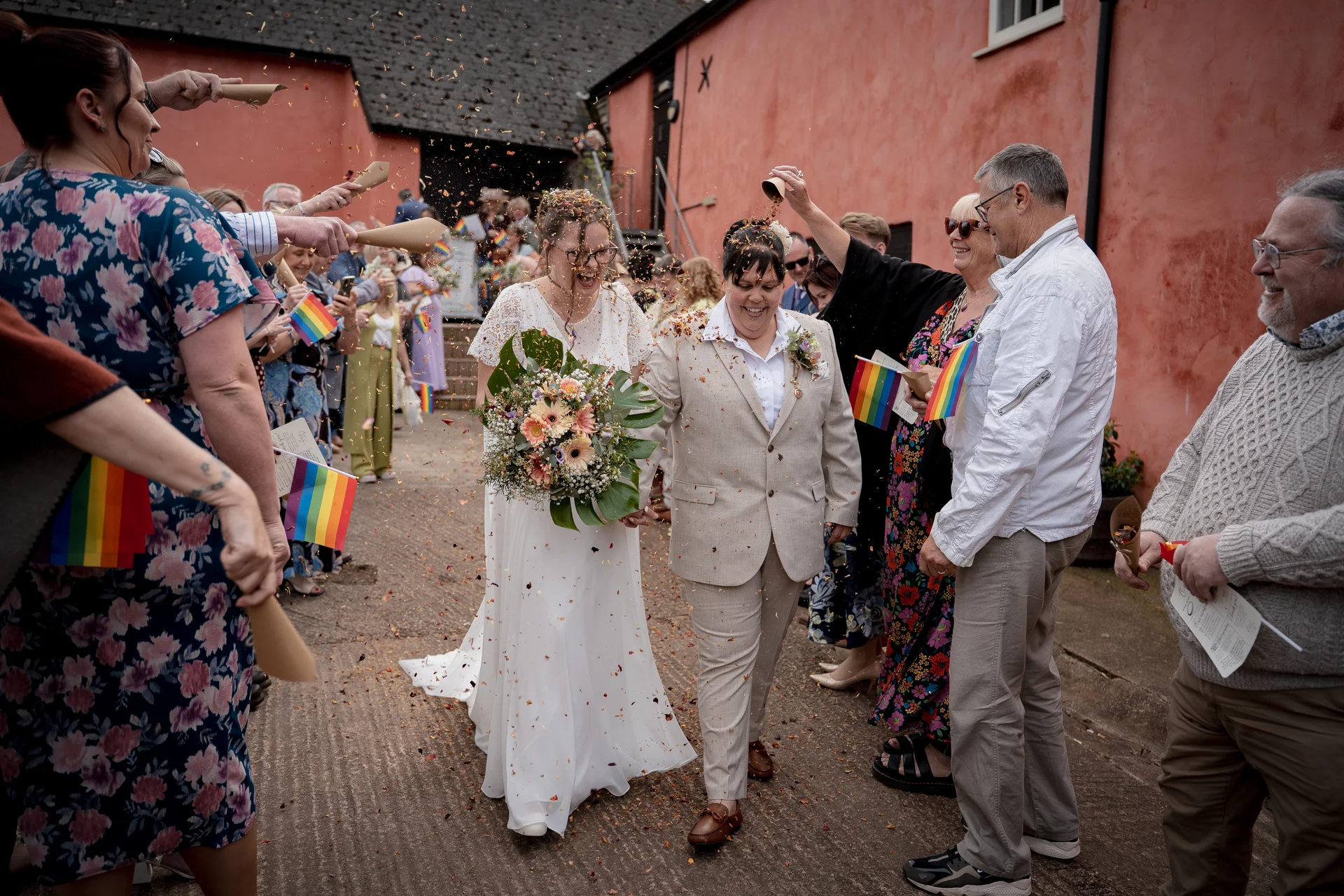 A same-sex couple joyfully walks through a shower of confetti held by cheering guests waving rainbow flags outside a rustic barn venue.