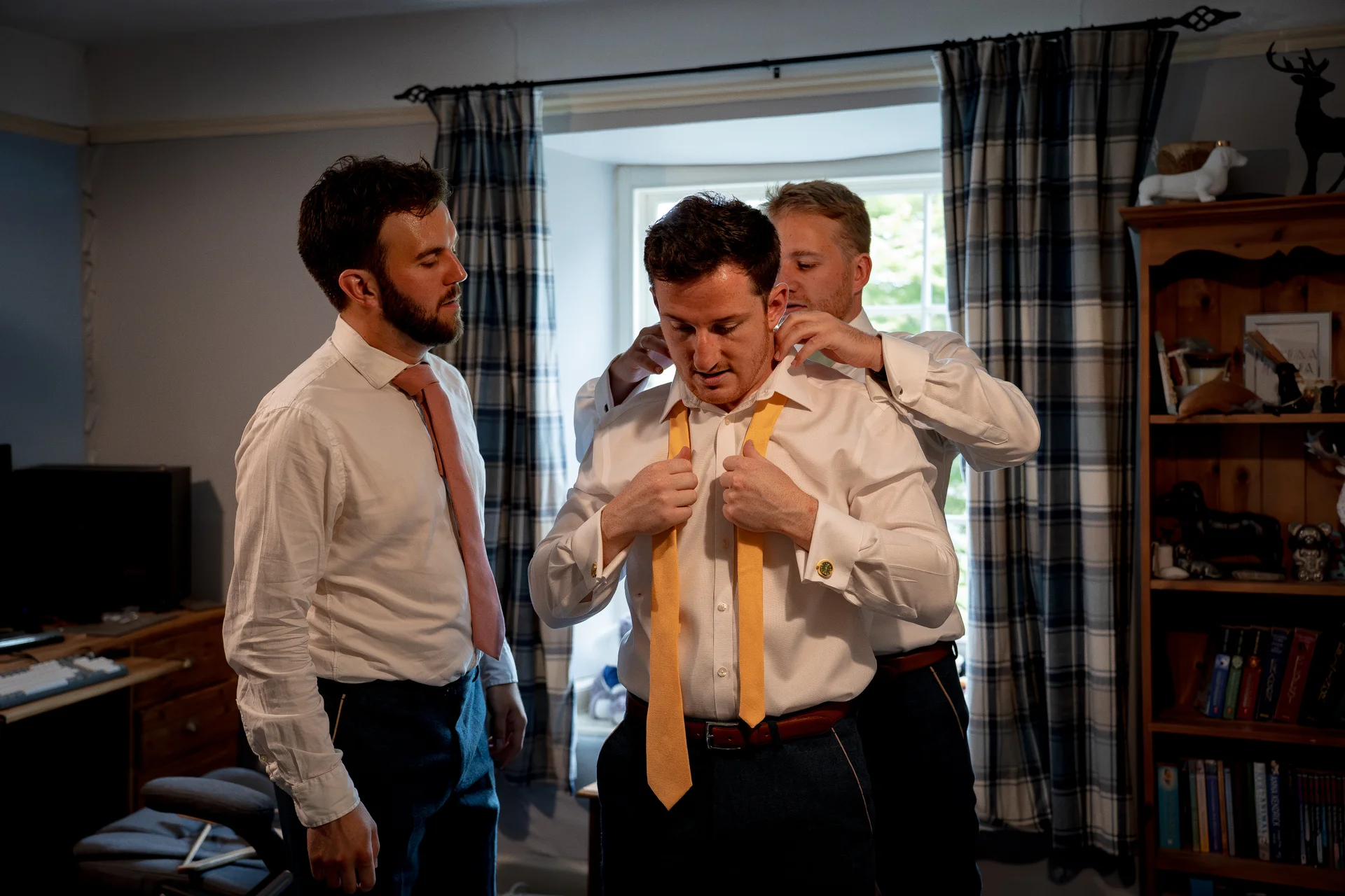 The groom is adjusting his yellow tie while getting ready with the help of his groomsmen in a room with a wooden bookshelf and plaid curtains.