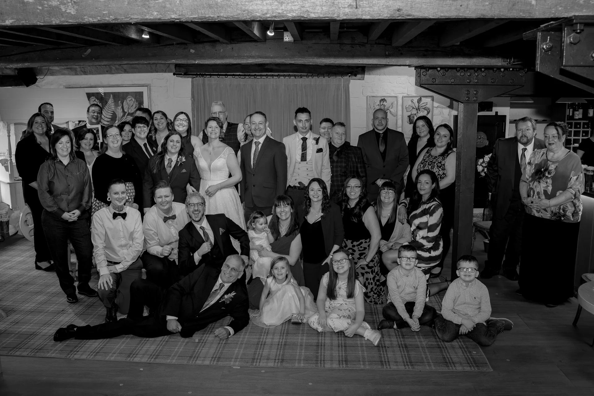 A large wedding group poses for a formal portrait inside a rustic barn with exposed wooden ceiling beams.