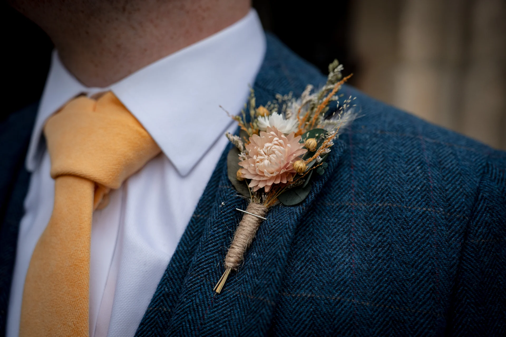 Close-up of a groom wearing a blue tweed suit with a white shirt and yellow tie, adorned with a dried floral buttonhole pinned to the lapel.