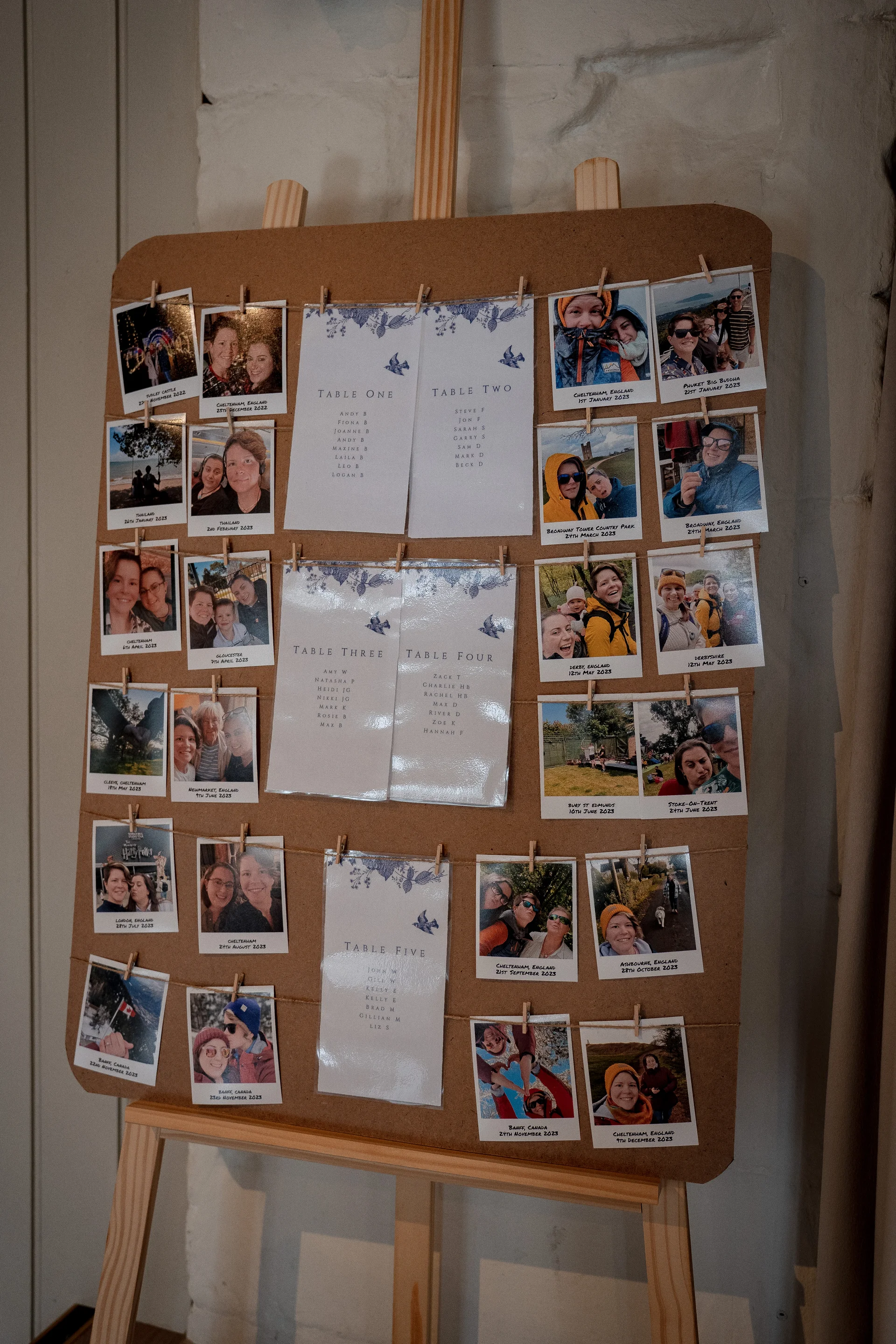 A rustic cork board displayed on a wooden easel features a wedding seating plan alongside numerous polaroid photographs clipped with miniature pegs.
