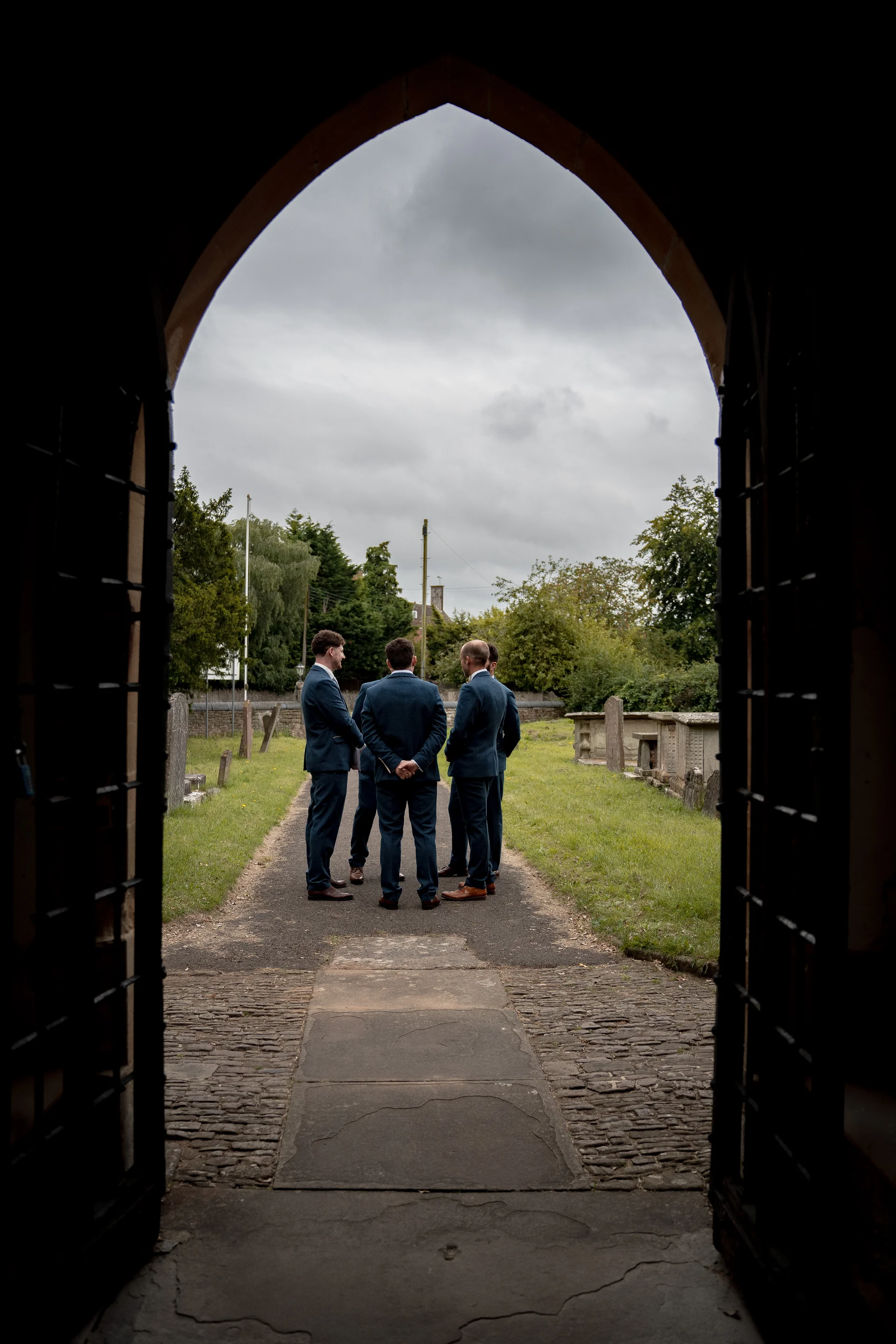 A group of groomsmen in matching blue tweed suits are framed through a stone archway, standing outside on a pathway near a church graveyard on an overcast day.