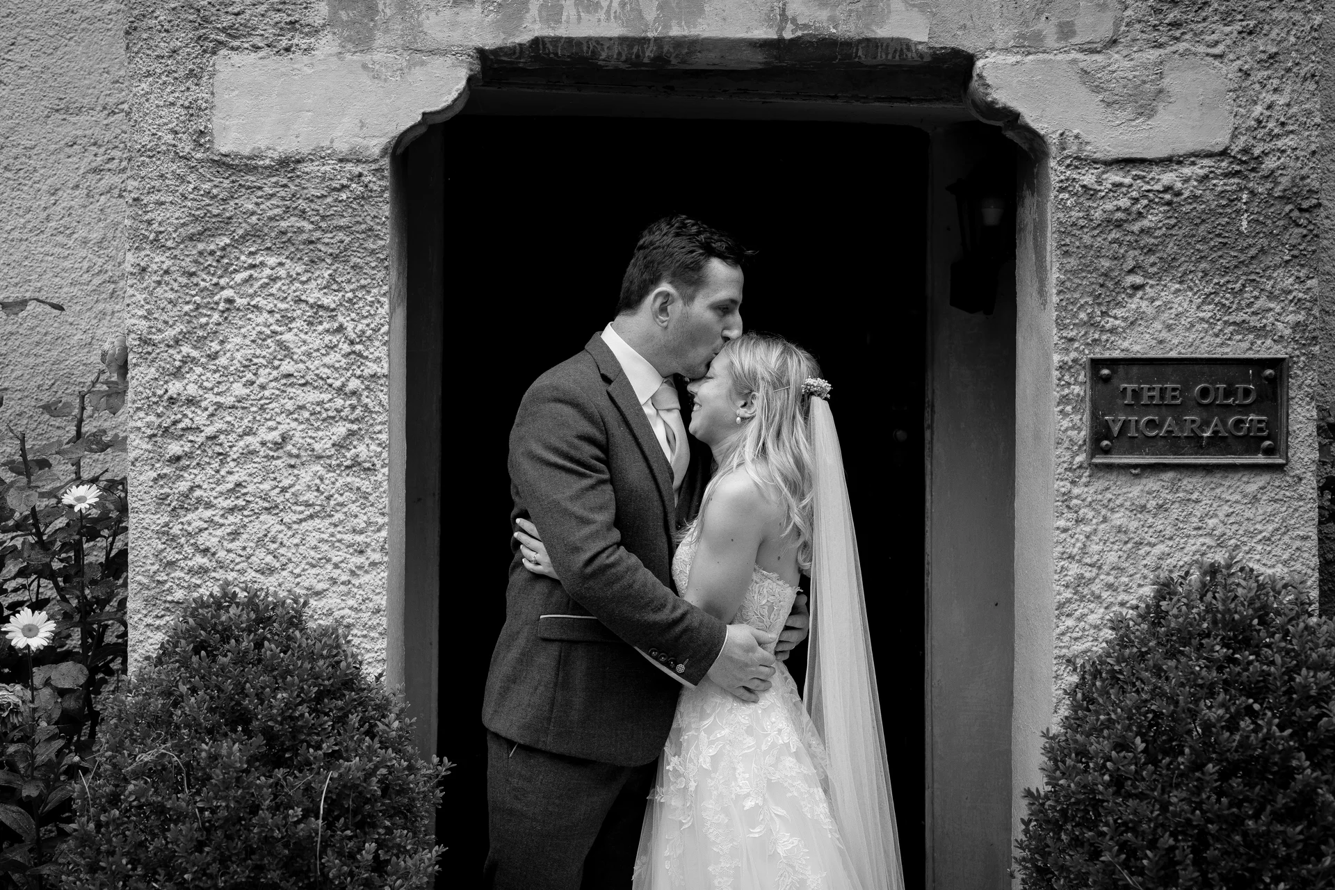A groom gently kisses his bride on the forehead as they stand framed within the stone doorway of a historic vicarage.