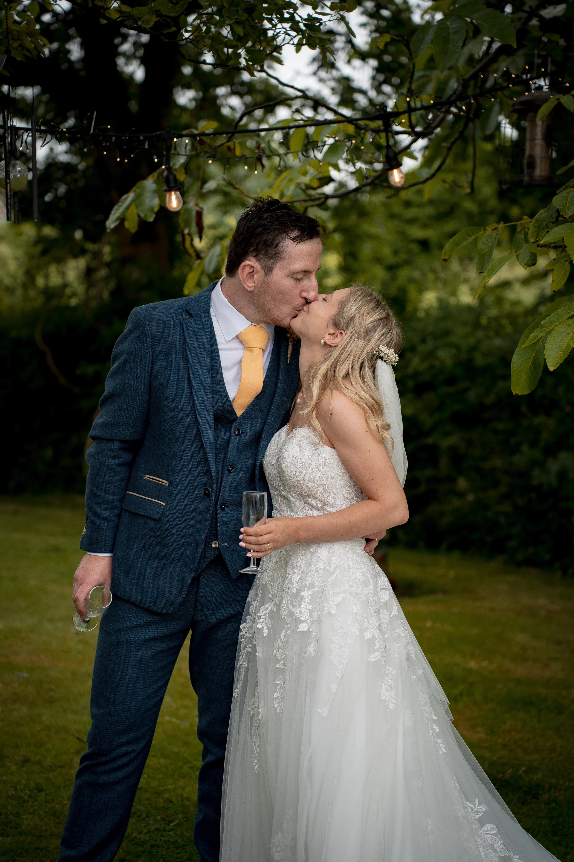 A newlywed couple share a tender kiss outdoors beneath a tree strung with fairy lights, holding champagne flutes during their garden reception.