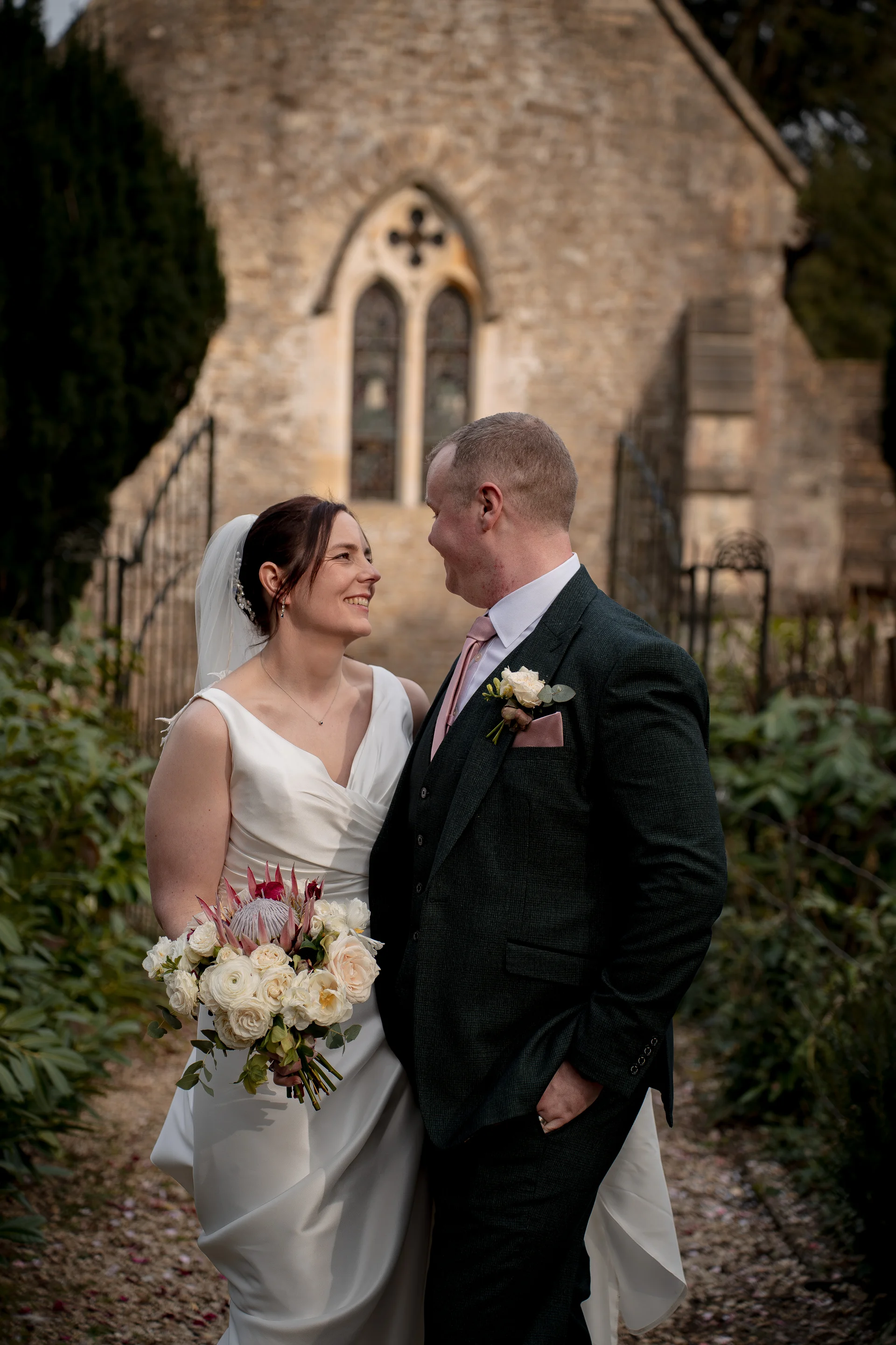 A smiling bride and groom gaze at each other lovingly outside a stone church, with the bride holding a textured bouquet containing a protea and roses.