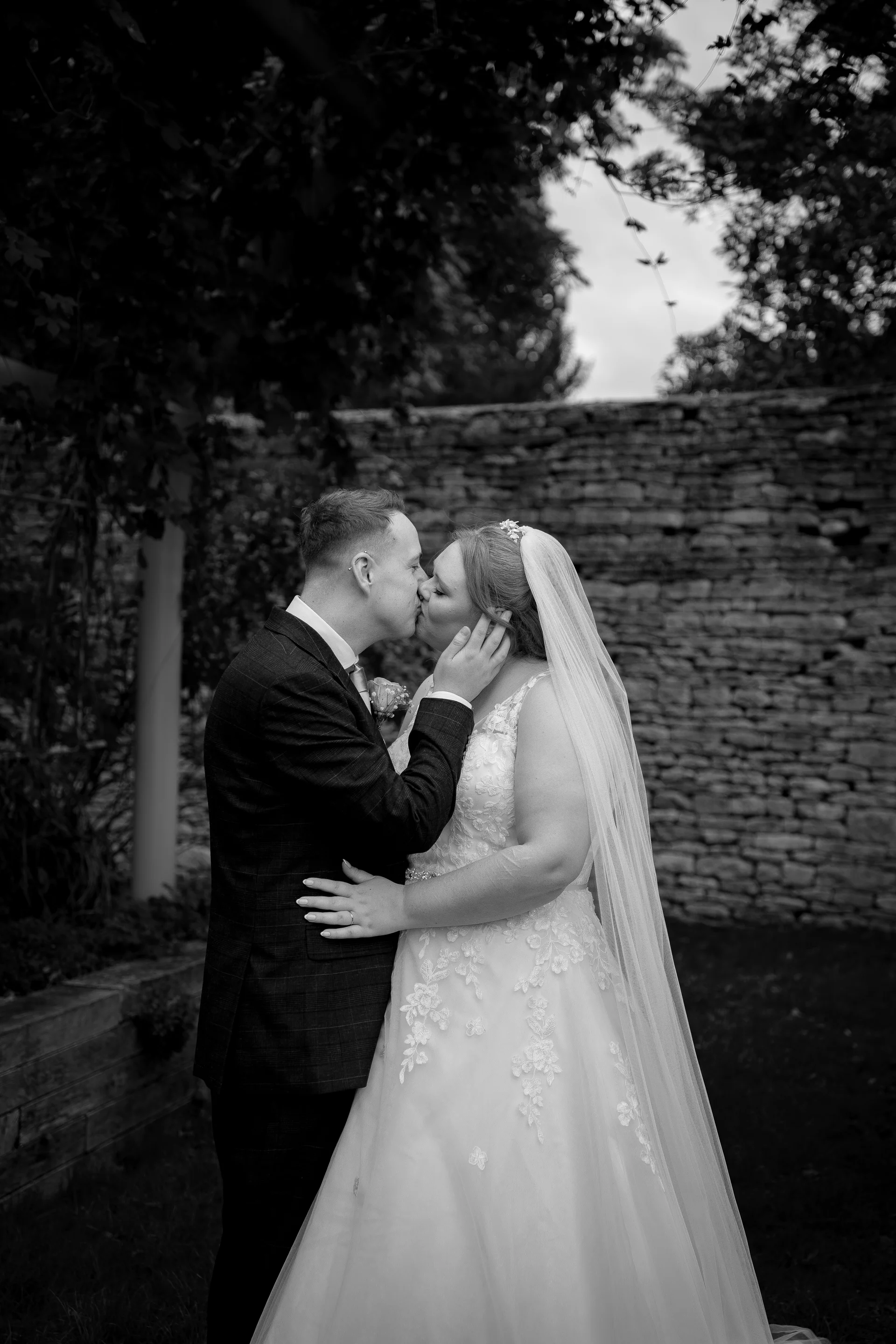 A newlywed couple share a tender kiss outdoors against a rustic stone wall, captured in a striking black and white photograph.