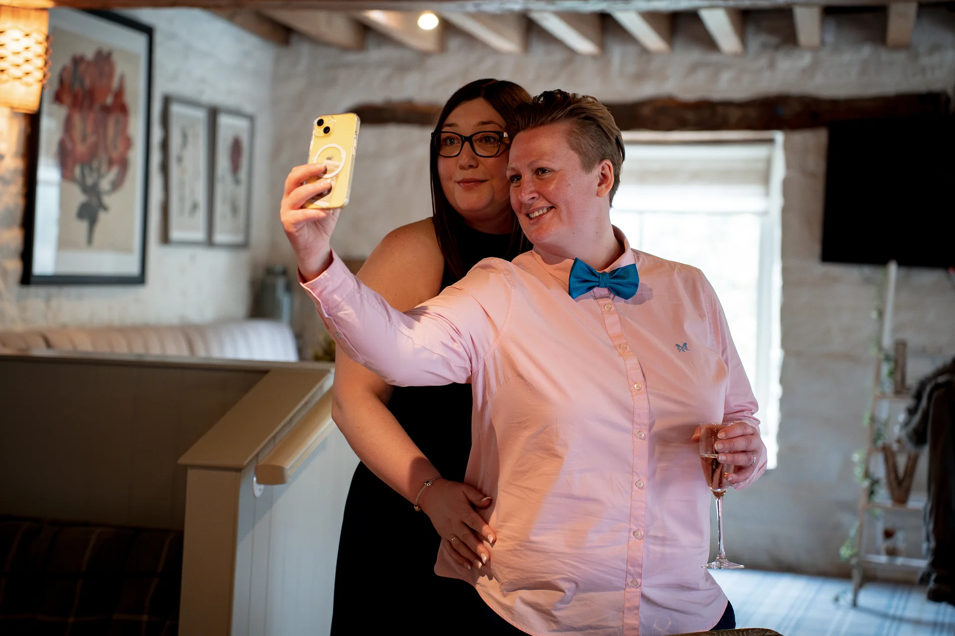 A same-sex couple smile brightly as one takes a selfie on a phone inside a rustic pub venue with exposed wooden beams.