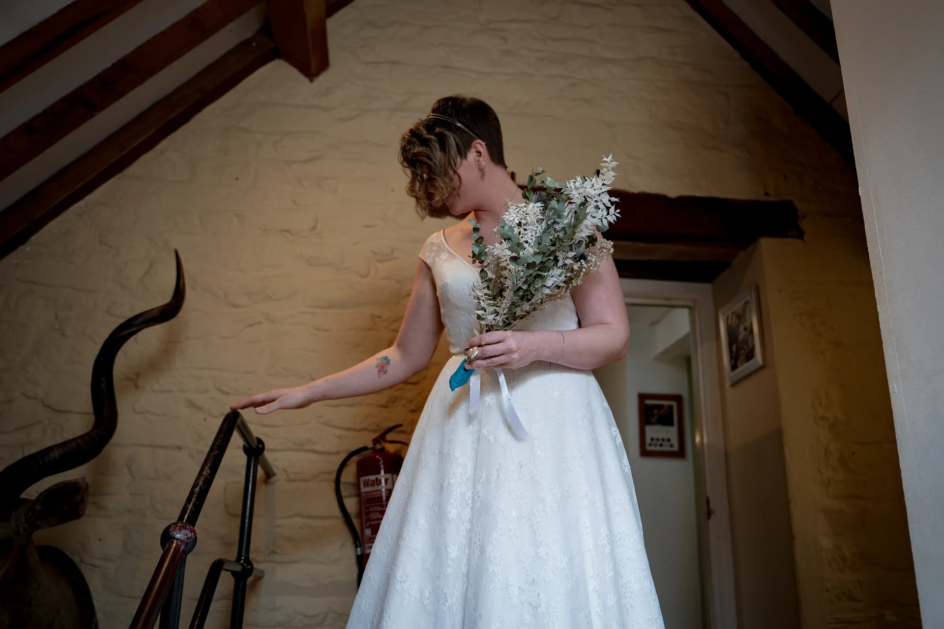 A short-haired bride in a lace a-line wedding dress holding a dried floral bouquet descends a stone staircase inside a rustic barn.