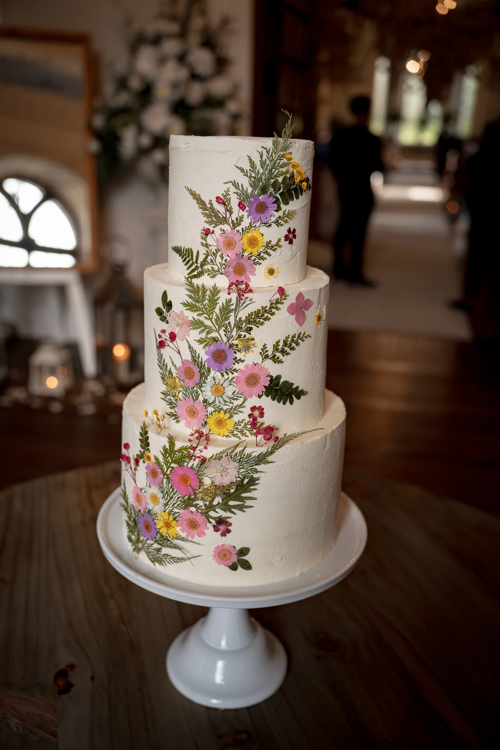 A three-tier white buttercream wedding cake is decorated with delicate pressed flowers and fern leaves cascading down its side, resting on a white cake stand.