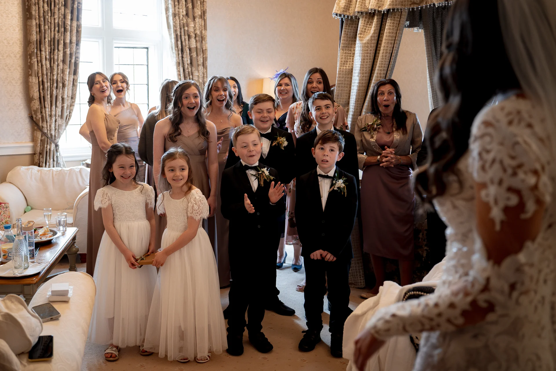 An emotional first look reveals the bride in her lace wedding dress to a delighted group of family, bridesmaids, flower girls, and page boys reacting with joy and surprise.