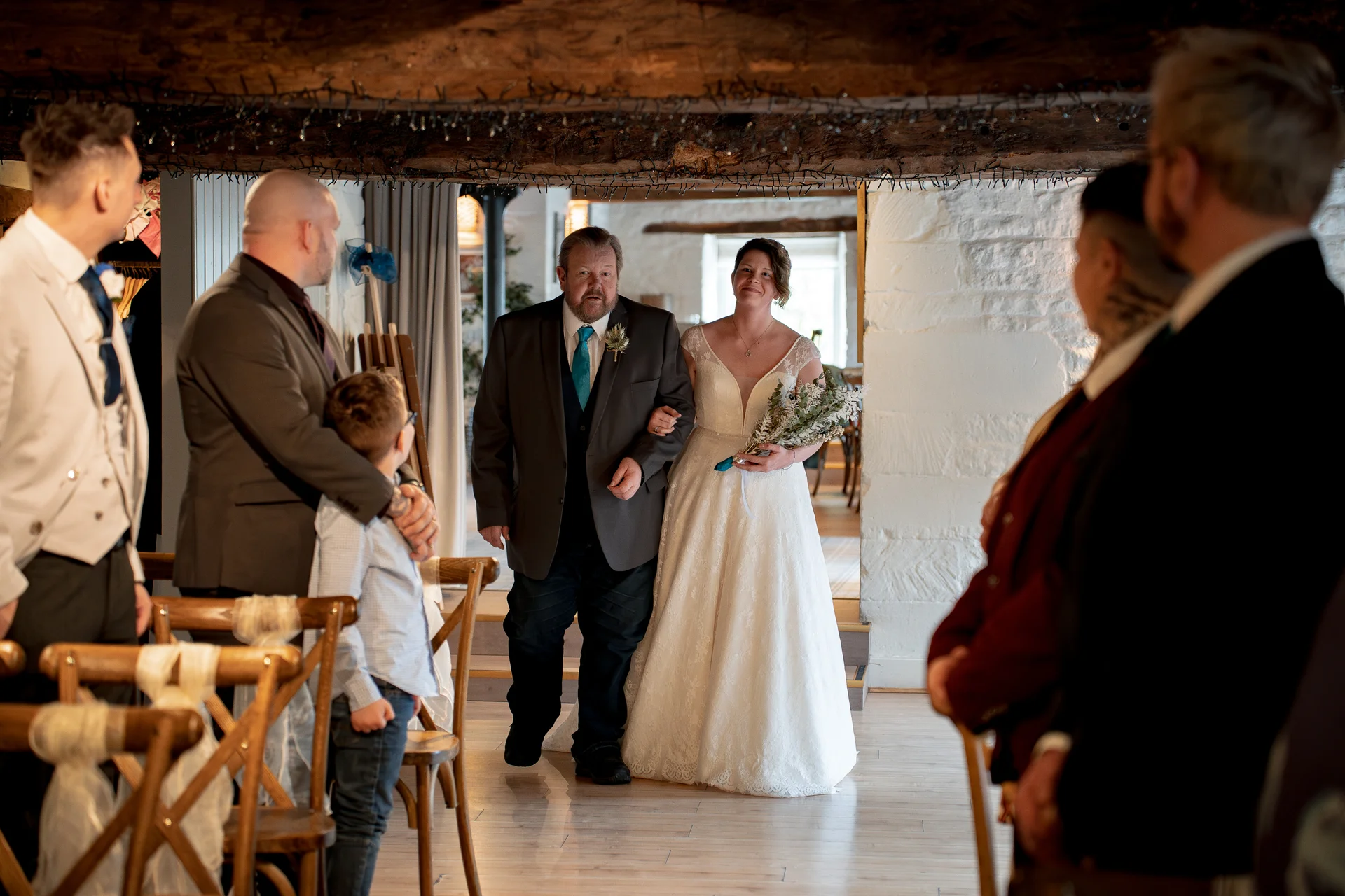 A father walks his smiling daughter down the aisle in a rustic stone barn venue, as guests watch from wooden cross-back chairs during the ceremony processional.
