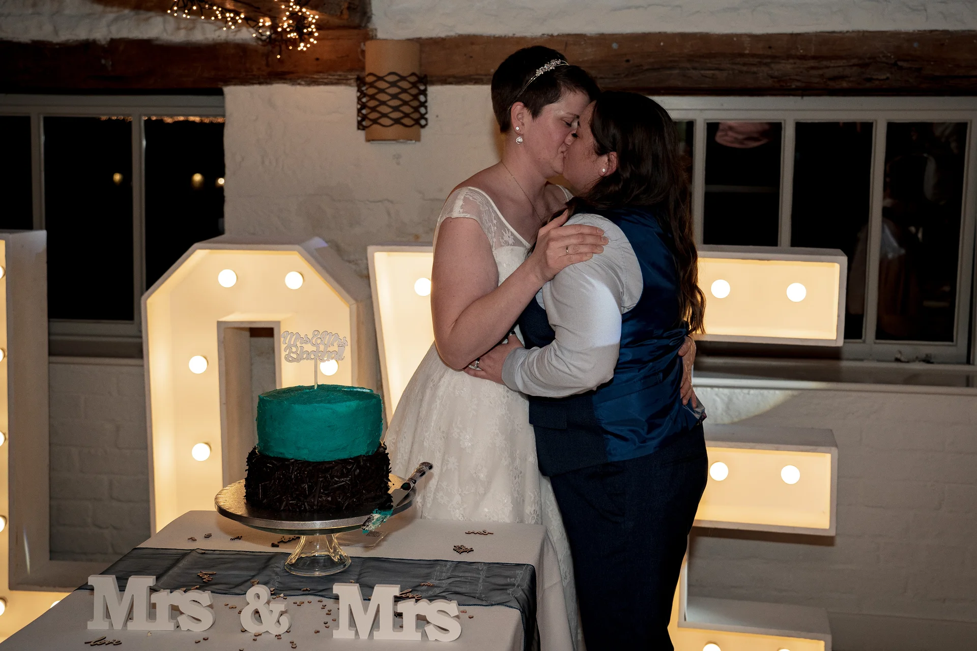 A newlywed same-sex couple share a kiss while cutting their teal and dark-tiered wedding cake in front of glowing marquee letters inside a rustic barn venue.