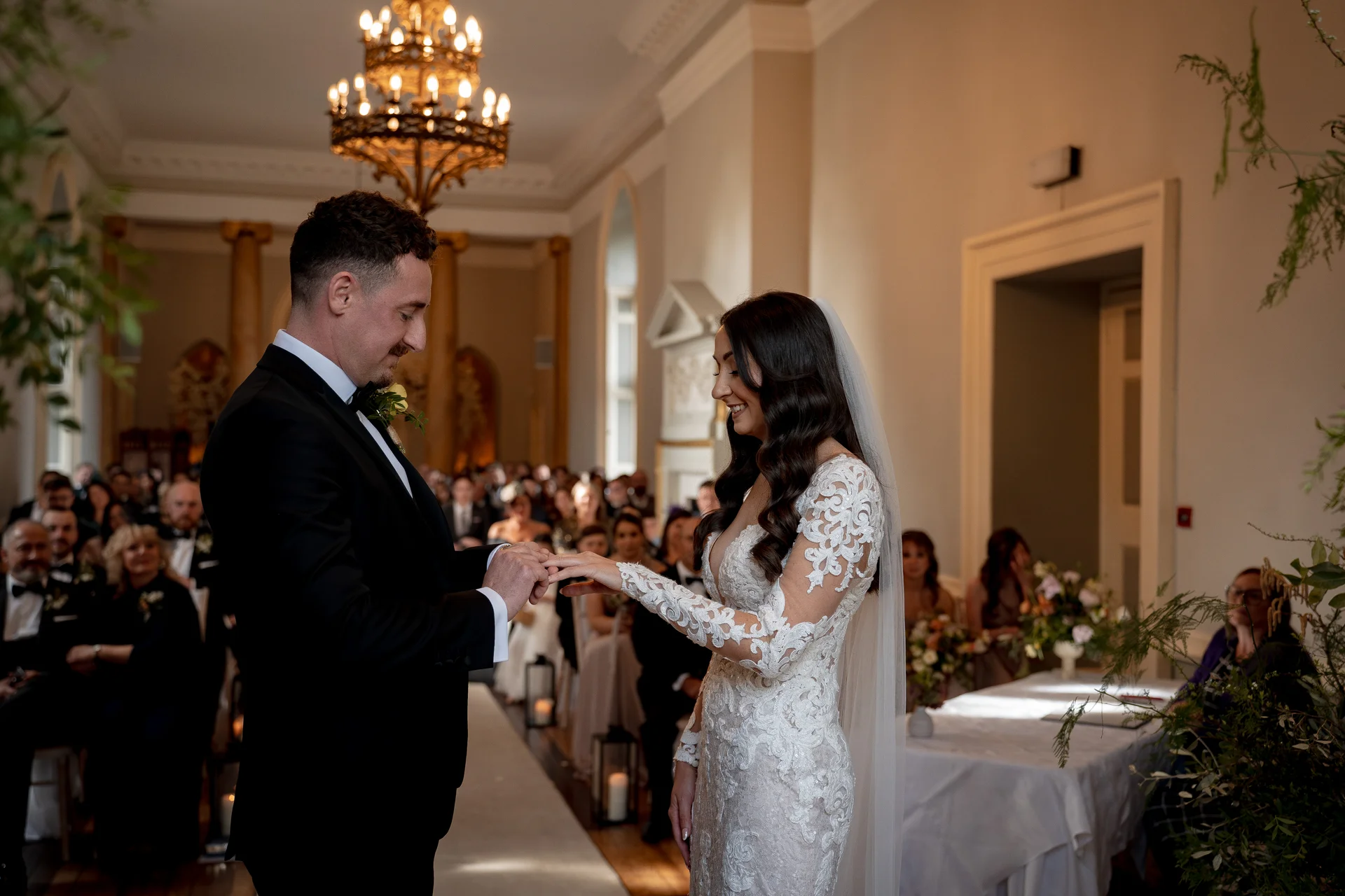 The groom gently slips a wedding ring onto the bride's finger during their indoor civil ceremony while seated guests watch on lovingly.