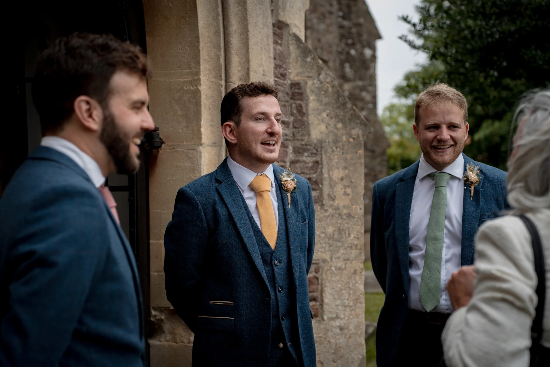 Three smiling groomsmen in blue tweed suits chat happily with a wedding guest outside a historic stone church during the drinks reception.