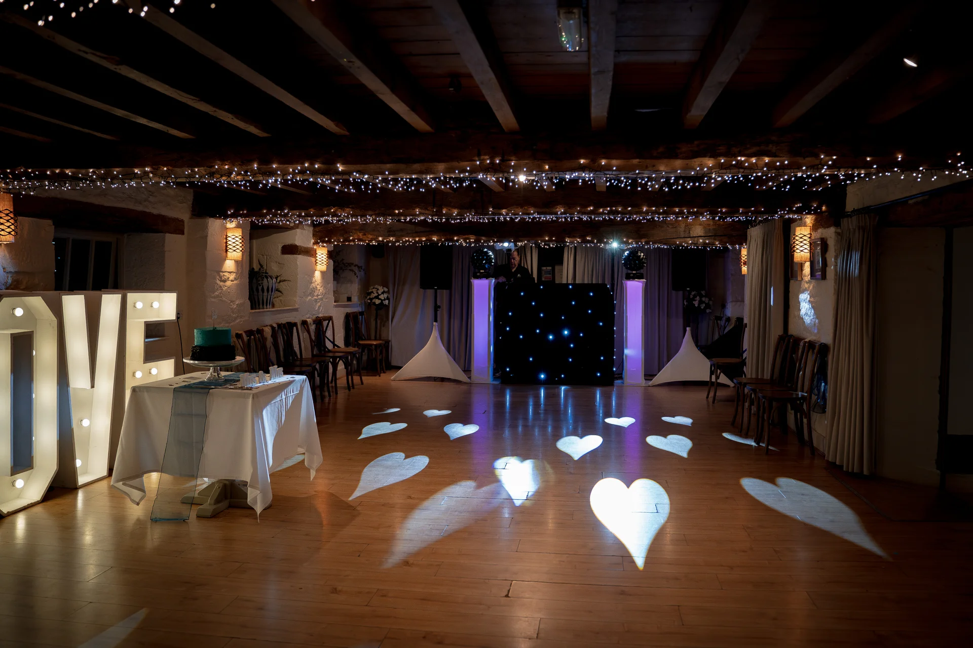 A rustic barn wedding venue at night featuring exposed wooden beams, glowing fairy light canopies, illuminated love letters, and heart-shaped light projections across the polished wooden dance floor.