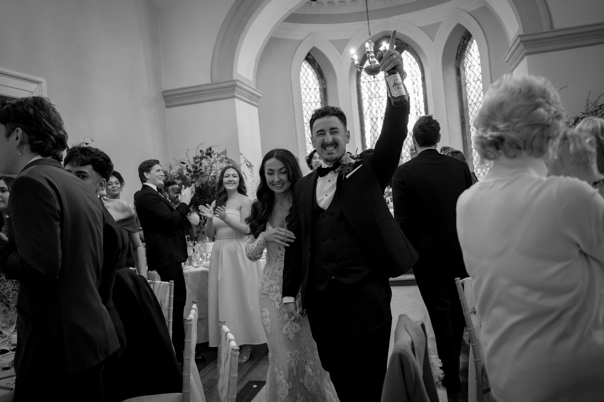 A newlywed groom joyfully raises a bottle in the air as he and his smiling bride walk hand in hand through an applauding crowd of wedding guests inside a historic venue.