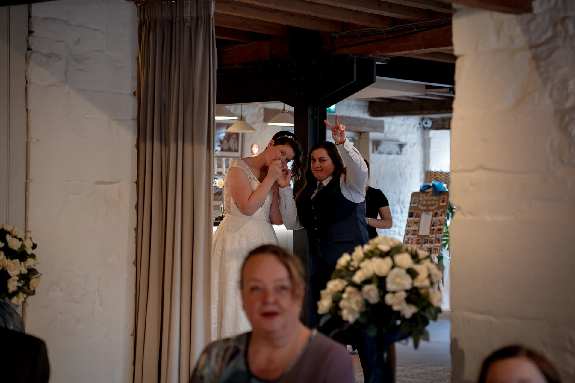 A same-sex couple shares a joyful, candid moment inside a rustic stone barn venue while guests are seated nearby.