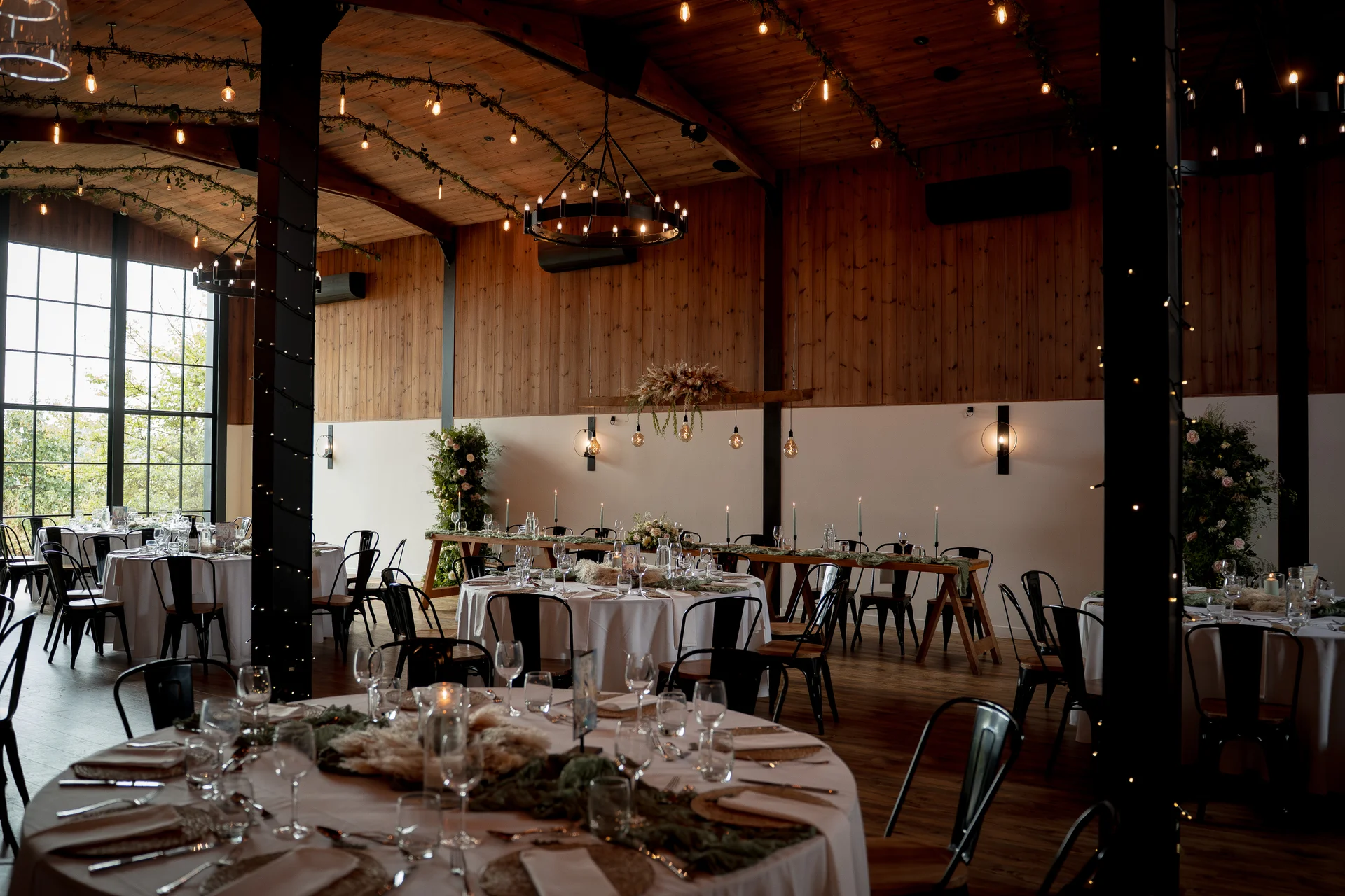 A spacious converted barn wedding venue featuring exposed wooden beams, large industrial windows, and round tables beautifully set with white linens and greenery.