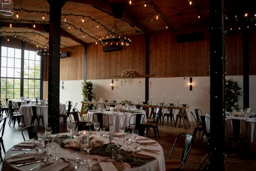 A spacious converted barn wedding venue featuring exposed wooden beams, large industrial windows, and round tables beautifully set with white linens and greenery.