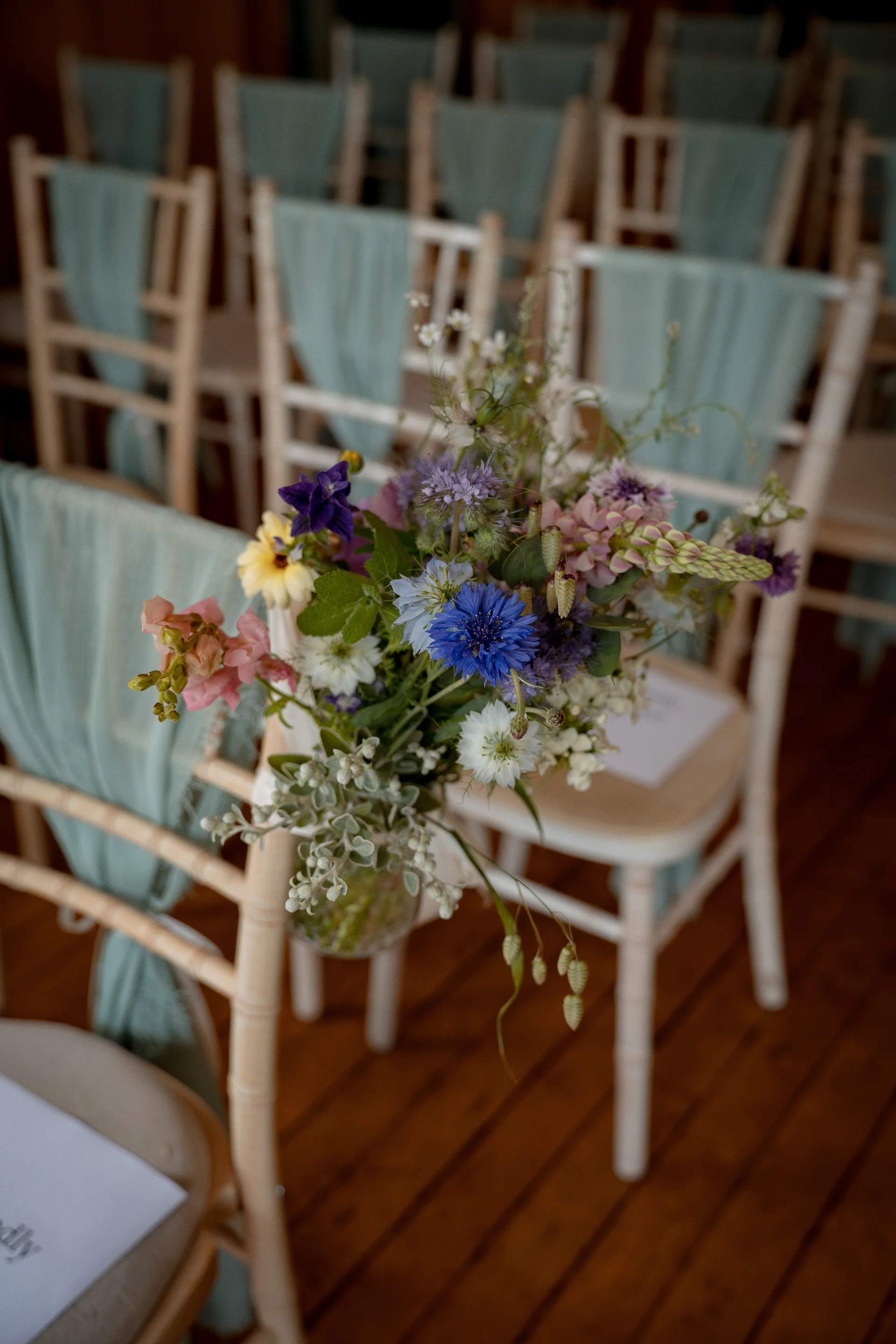 A rustic wedding aisle features wooden chairs adorned with wild blue, yellow, and purple flowers and pale sage green drapes.