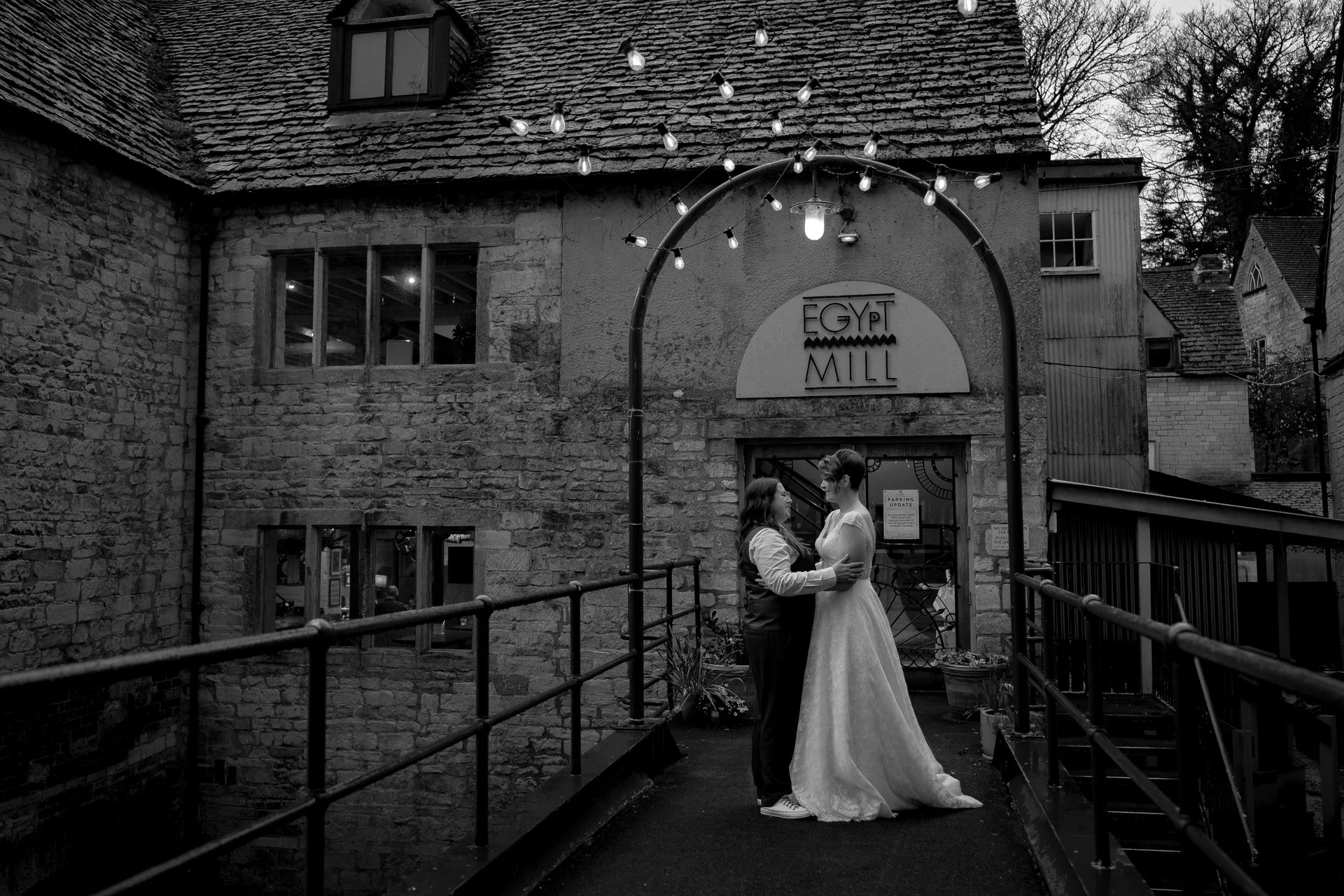 A same-sex couple shares a tender embrace on a stone bridge outside a rustic barn venue illuminated by string lights.