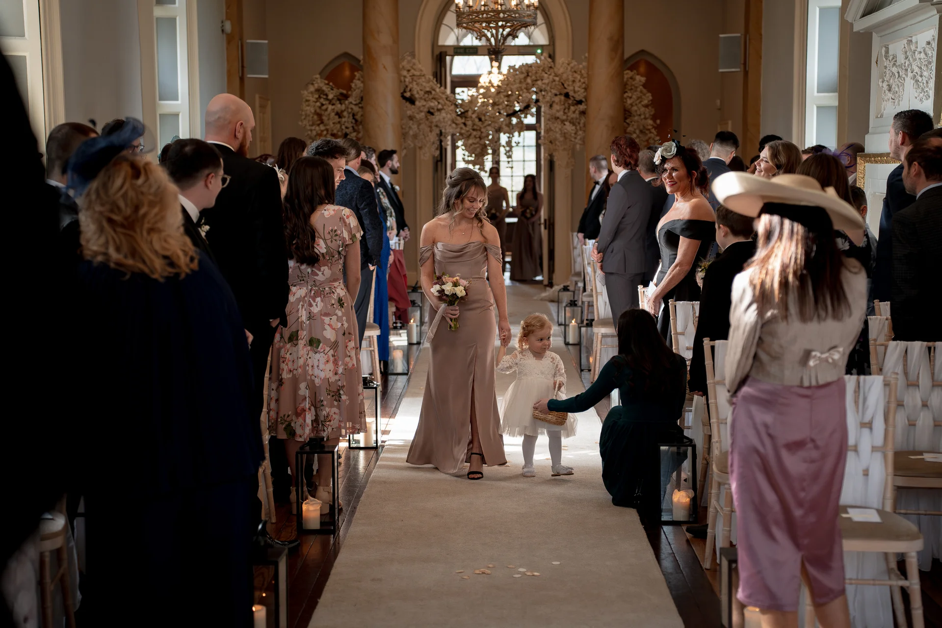 A flower girl holding hands with a bridesmaid wearing a sleek taupe gown walks down the aisle during a grand indoor wedding ceremony.