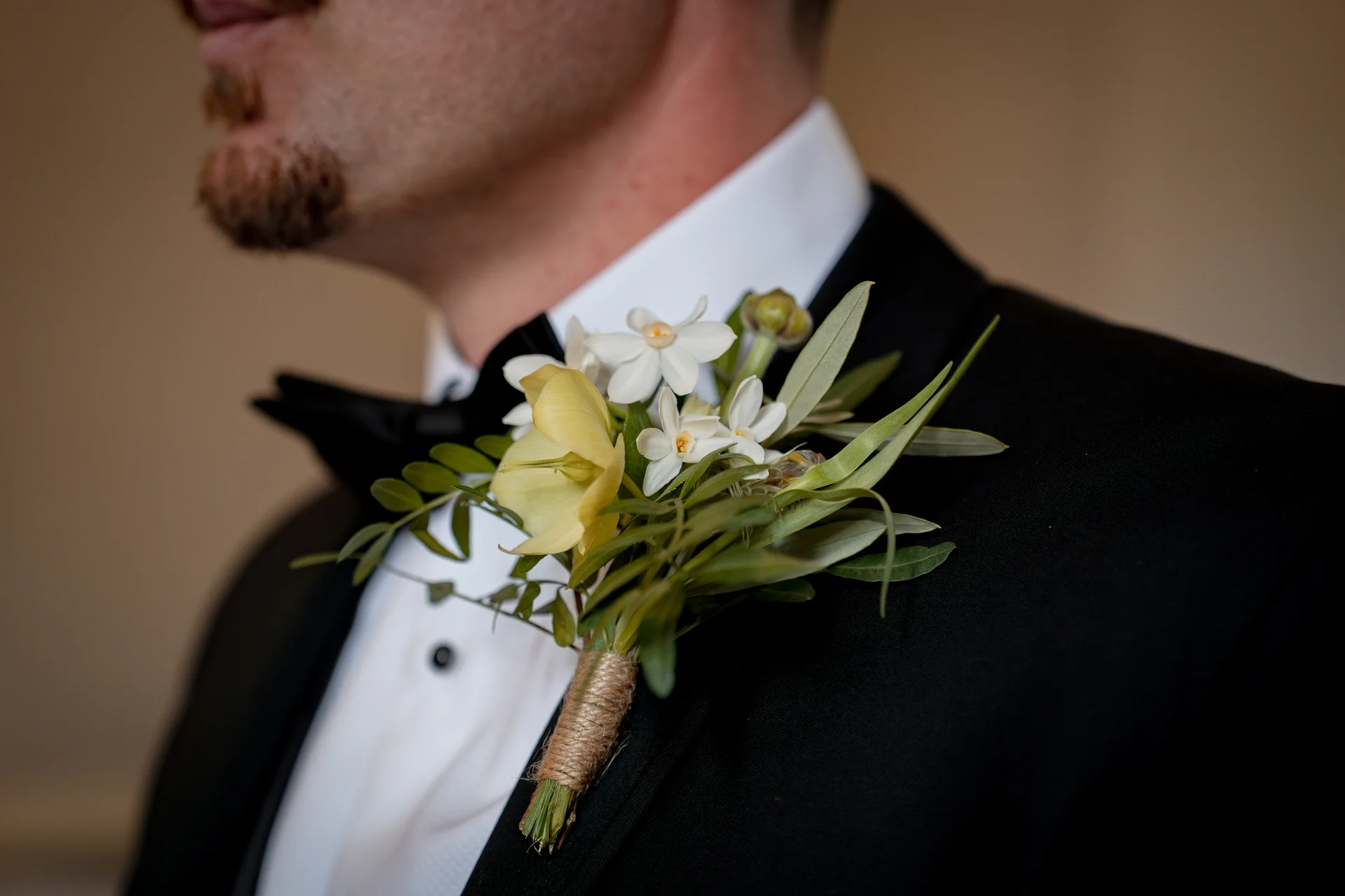 A close-up shot of a groom wearing a classic black tuxedo and bowtie, adorned with a delicate floral buttonhole featuring white and yellow flowers.