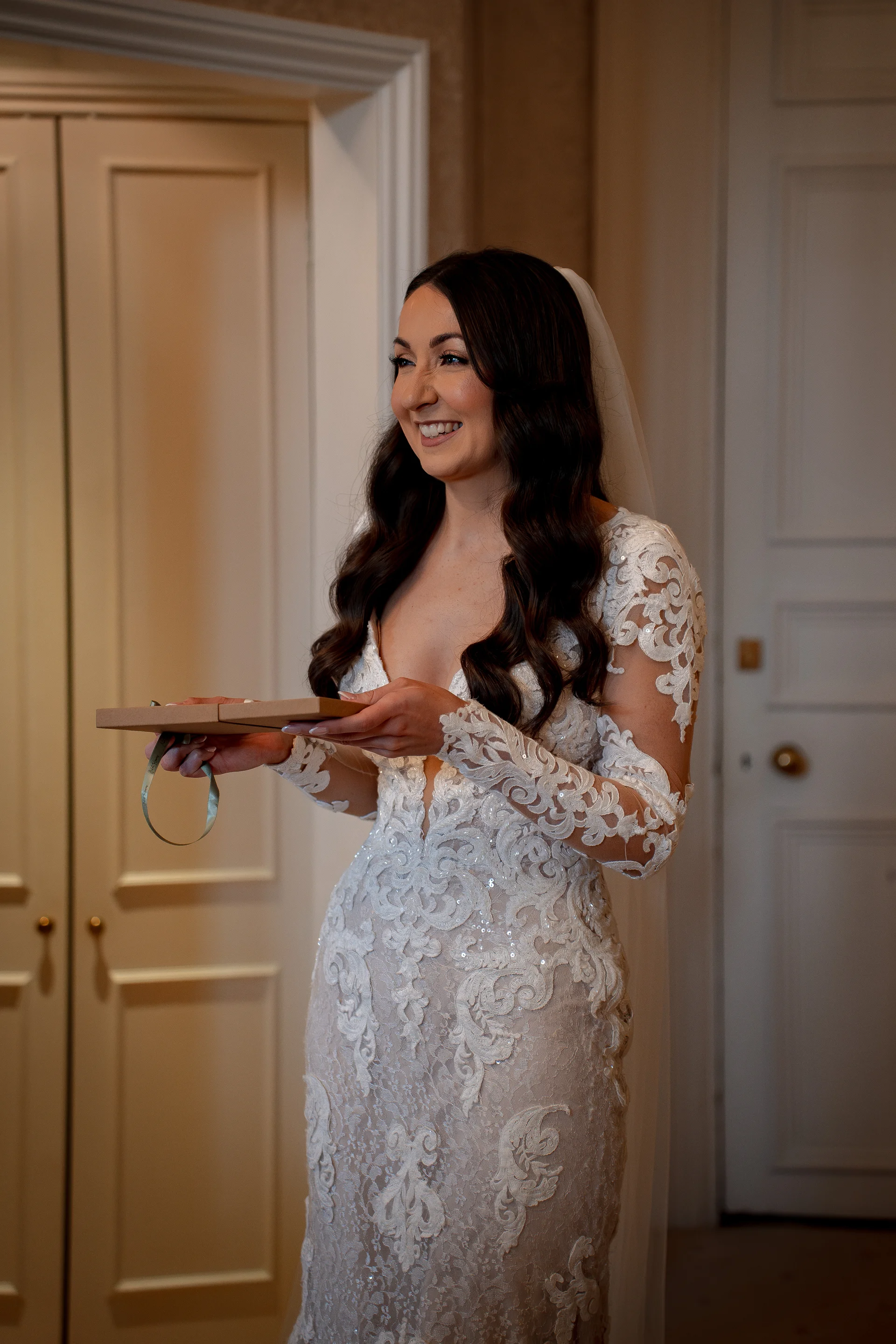 A smiling bride wearing an intricate lace long-sleeve wedding dress stands indoors, holding a small wrapped gift during bridal preparations.