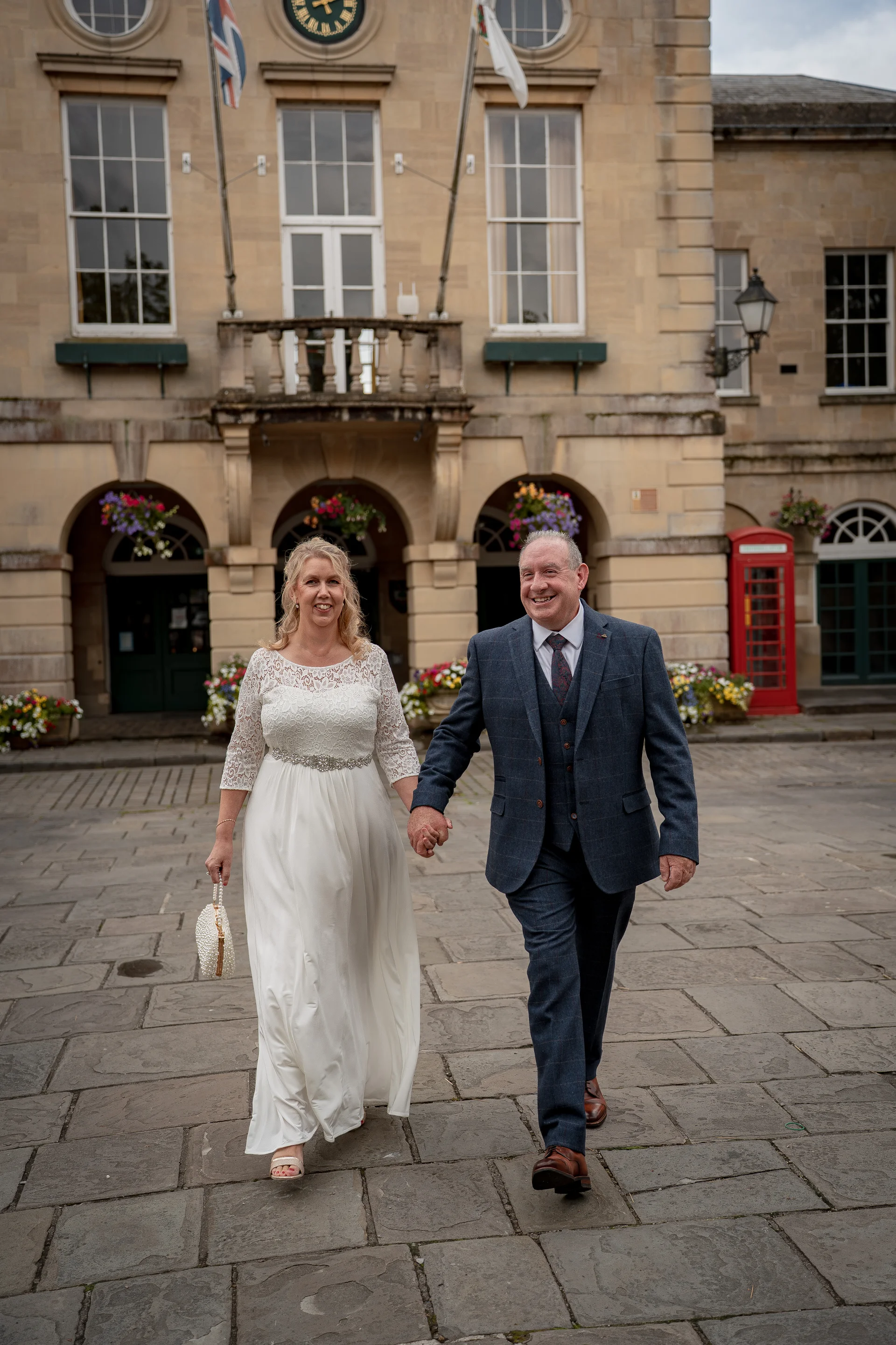 A mature bride and groom walk hand in hand across a historic cobbled courtyard in front of a grand stone town hall.