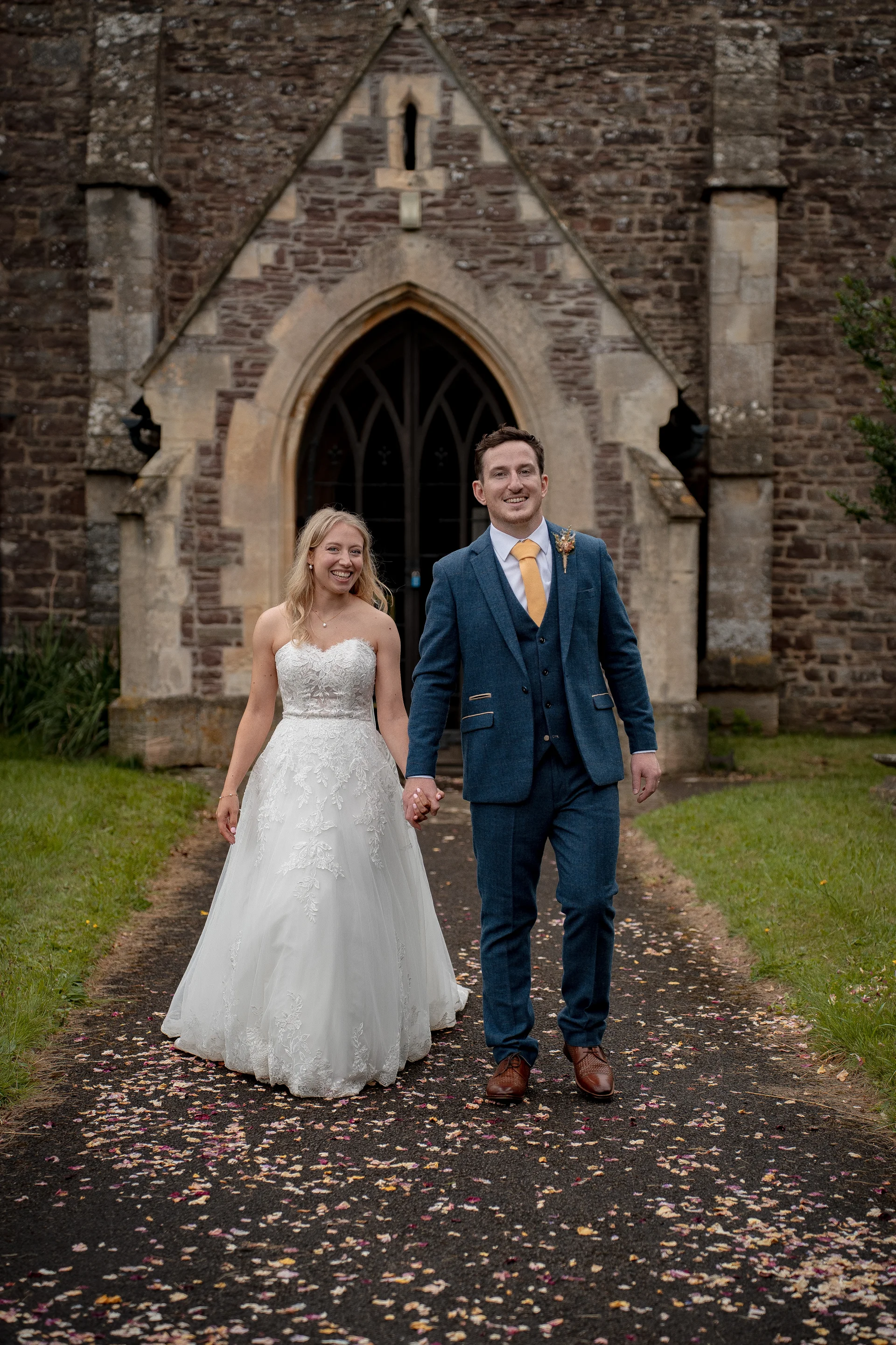 A smiling bride and groom walk hand in hand down a path scattered with petals outside a historic stone church.