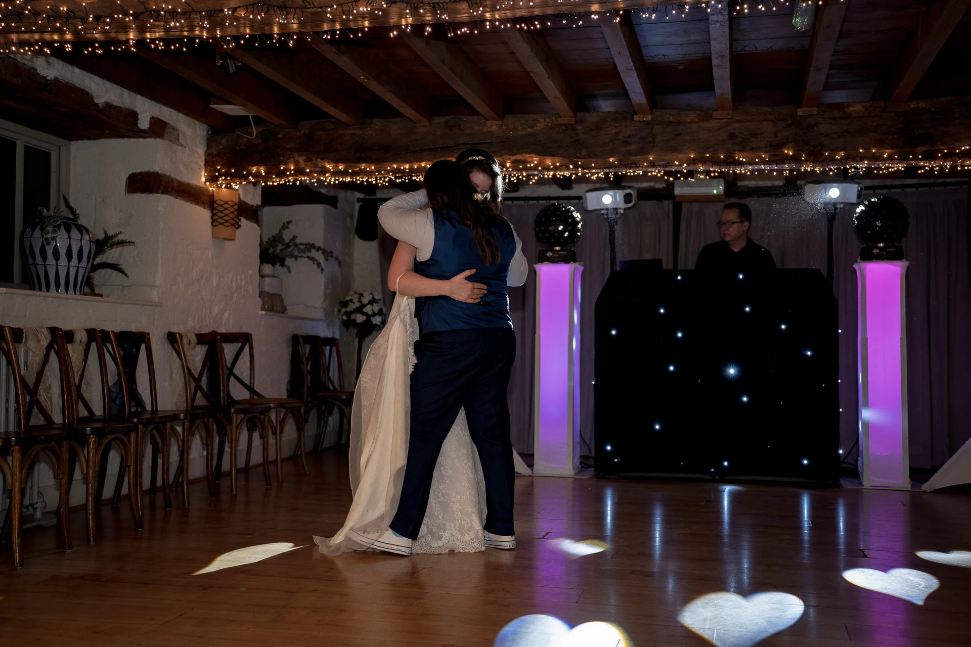 A same sex couple shares an embrace during their first dance under a canopy of fairy lights in a rustic barn venue with a DJ booth in the background.